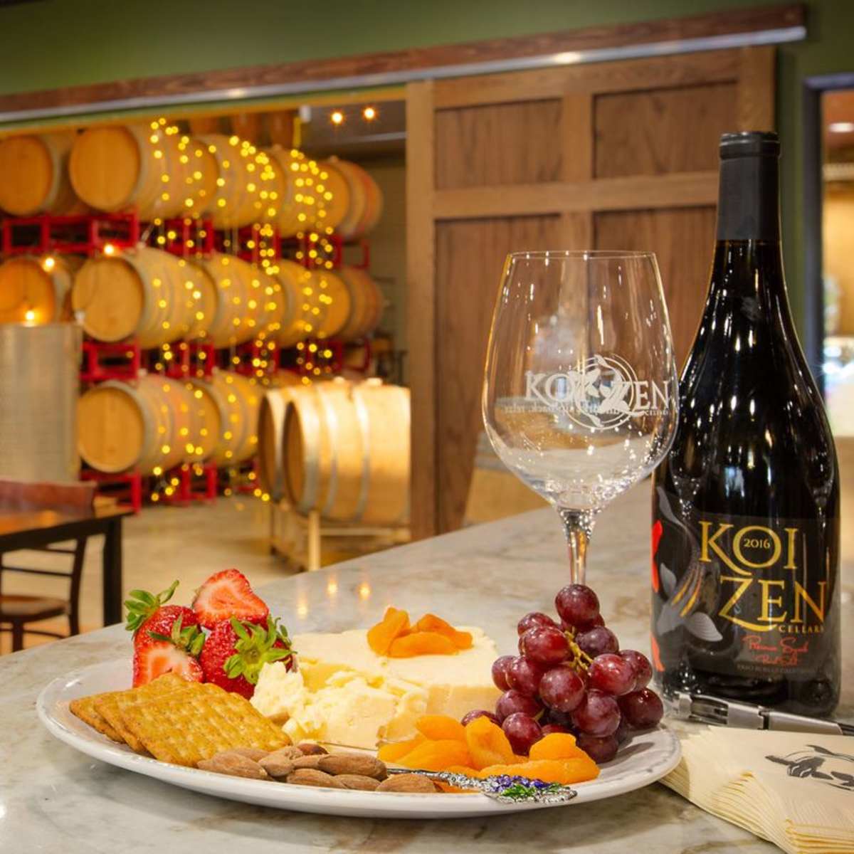 A wine tasting room with a plate of cheese, fruits, and nuts in the foreground, and wooden wine barrels in the background.
