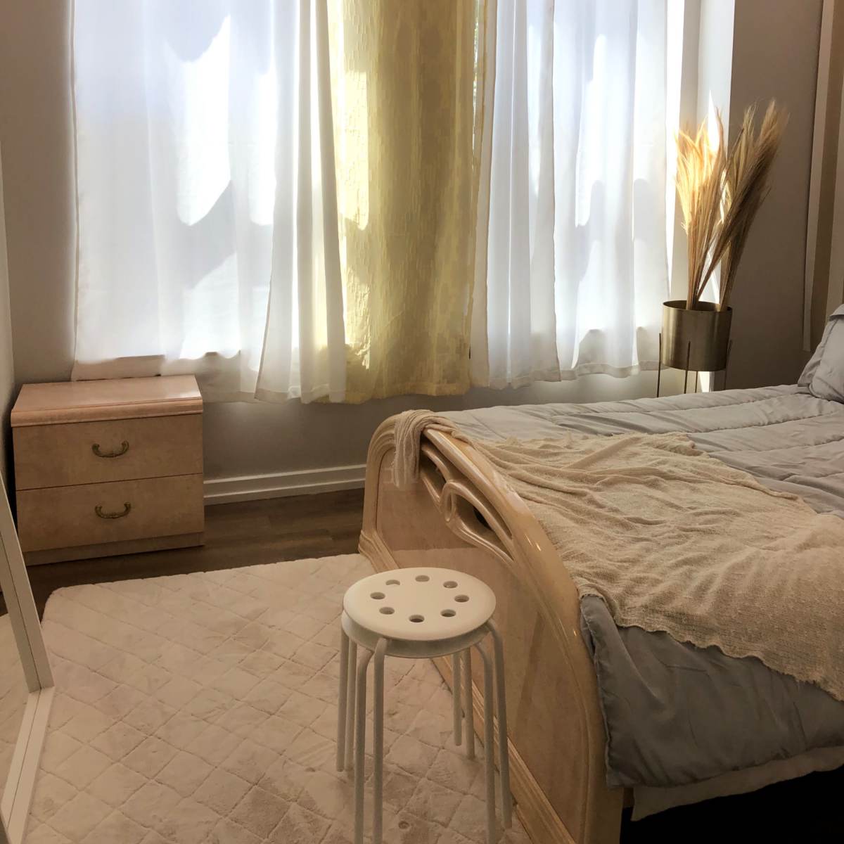 A bedroom features a bed with a soft blanket, a nightstand, and large curtains letting in filtered light.