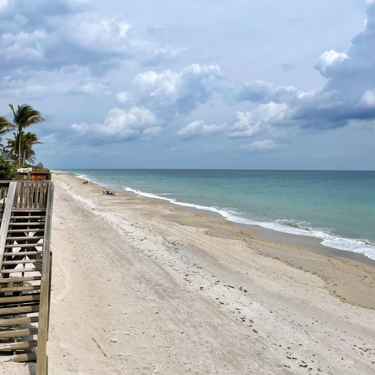 A sandy beach with a wooden staircase leading down from a grassy area, under a cloudy sky.
