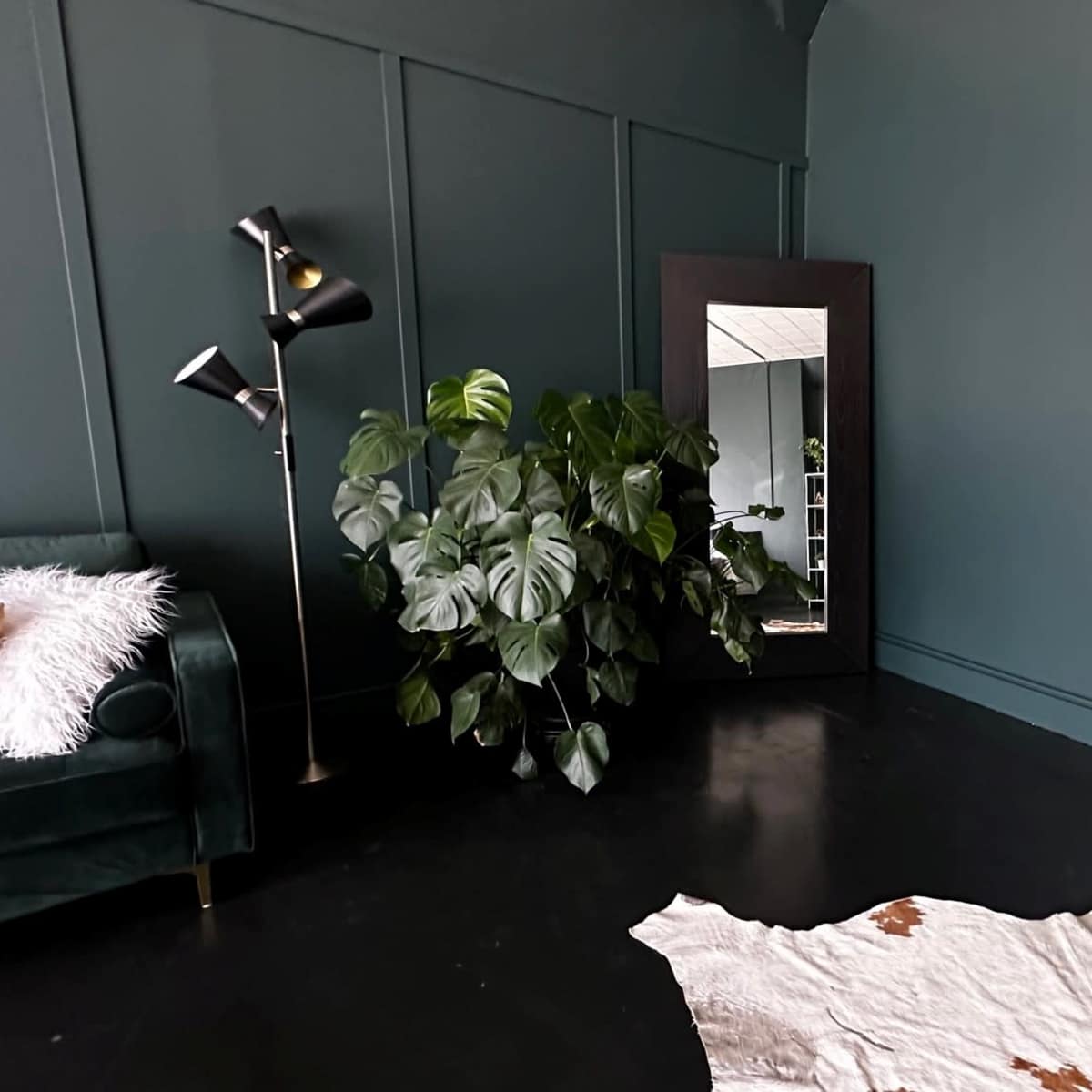A room with deep green walls, a large mirror, a leafy plant, a floor lamp, a green sofa, and a cowhide rug on a dark floor.