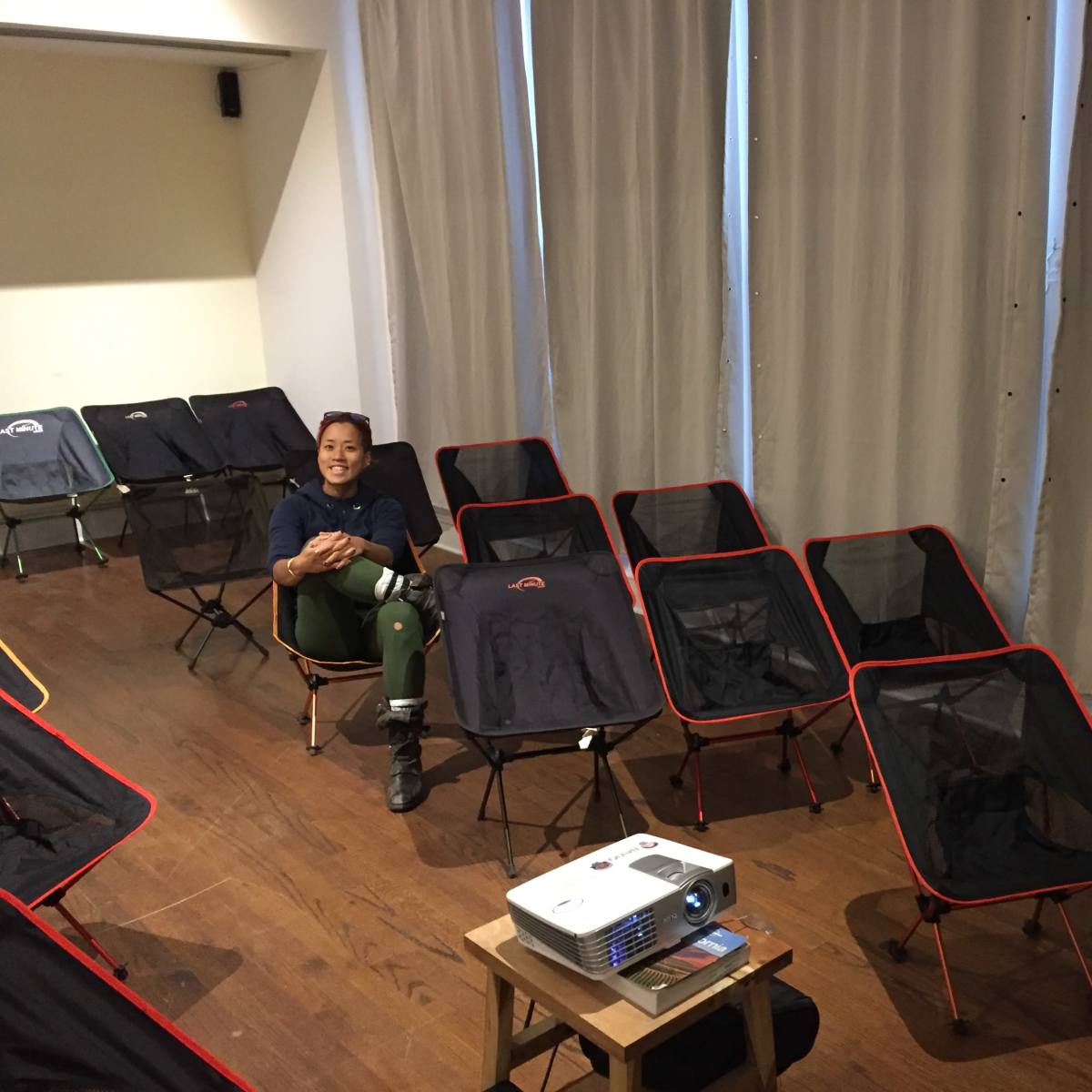 A person sits among several folding chairs in a room with curtains and a projector placed on a small table.