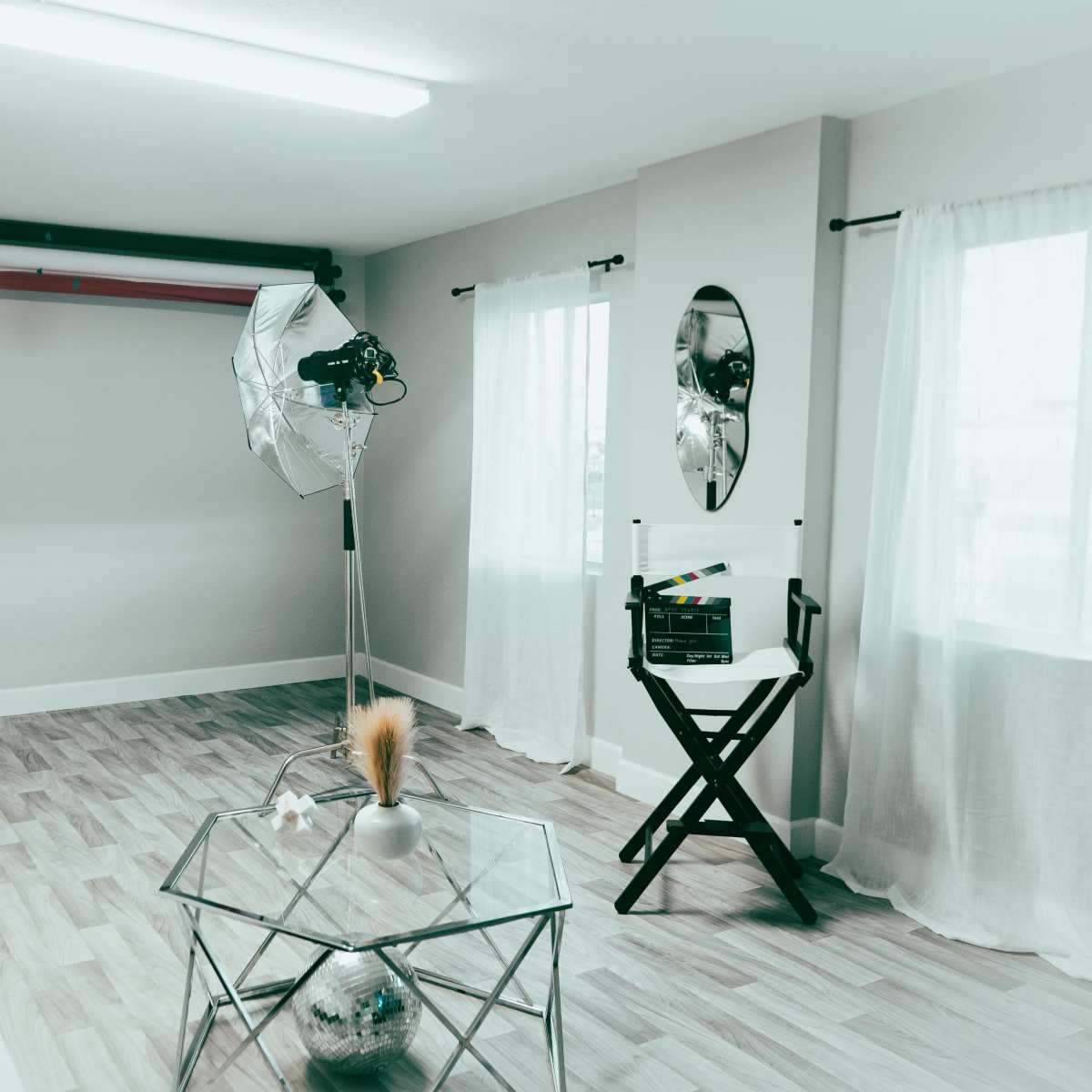 A bright photography studio features a white couch, a standing mirror, and two large lights set up beside a director's chair and a glass coffee table.