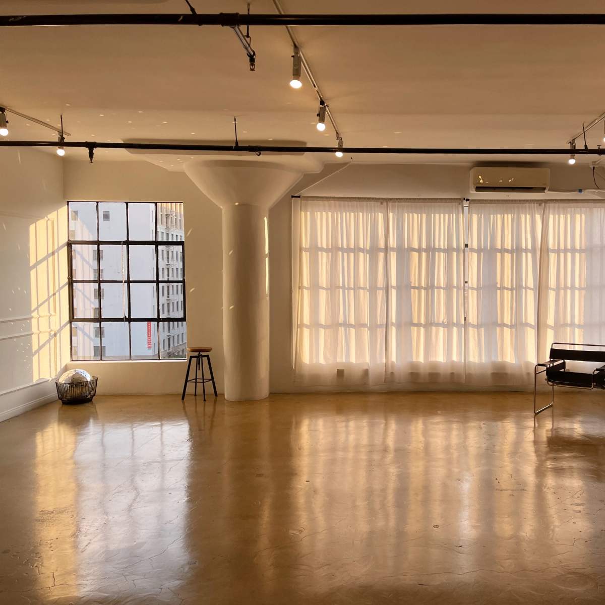 A spacious, well-lit studio with large windows covered by sheer curtains, a brown sofa, and minimal furnishings.