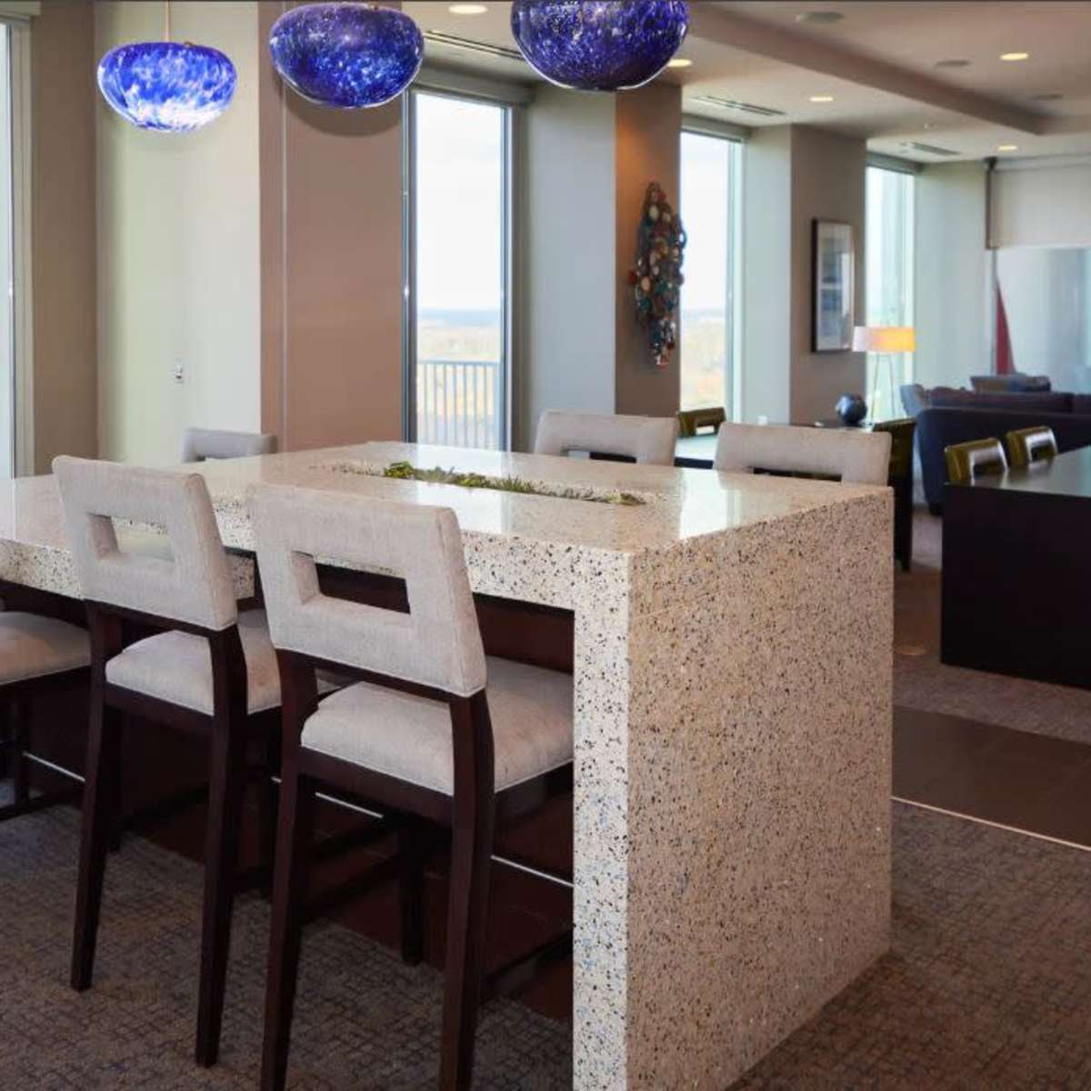 A modern kitchen and dining area features a large stone island with bar seating, complemented by pendant lighting and large windows offering a view of the outside.