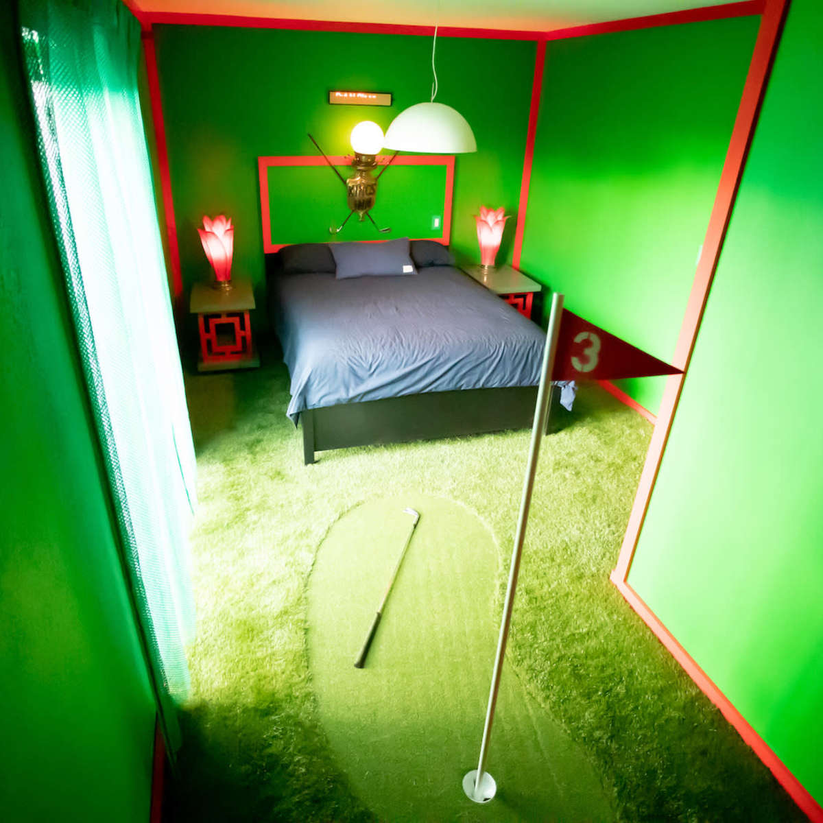 A small, brightly colored bedroom with green walls, a bed with blue bedding, and a putting green on the floor.