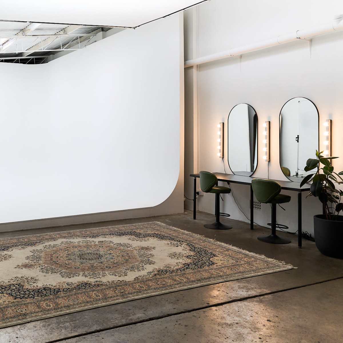 A spacious photography studio featuring a seamless white backdrop, a vintage rug on the floor, a vanity with illuminated mirrors, and a potted plant.