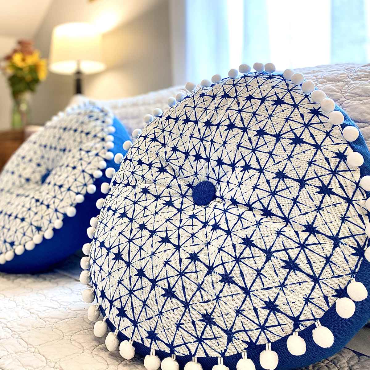 Two round blue and white decorative pillows with pom-pom edges rest on a neatly made bed in a softly lit room.