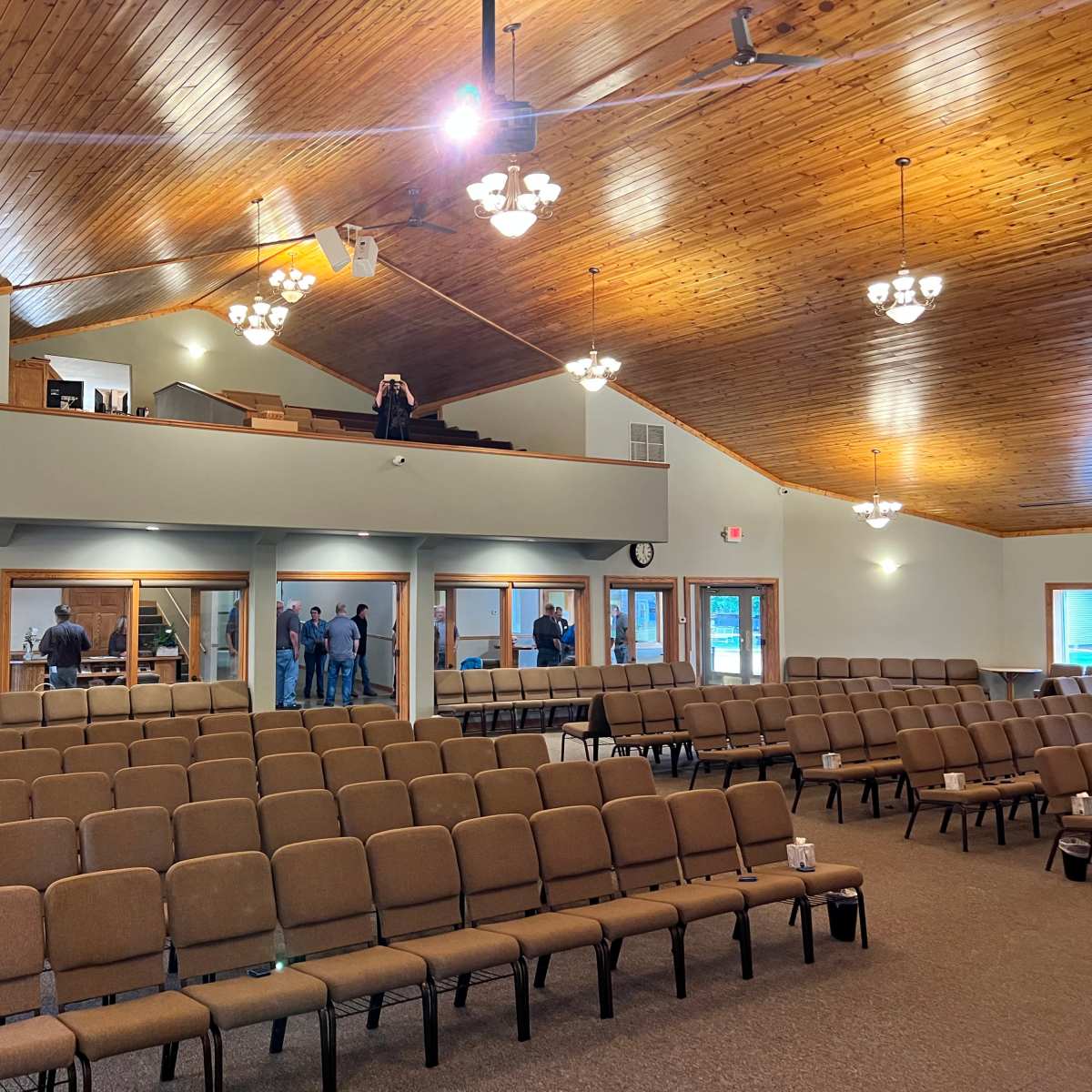 A spacious room with rows of brown chairs, wooden ceilings, and a small group of people gathered near the back.