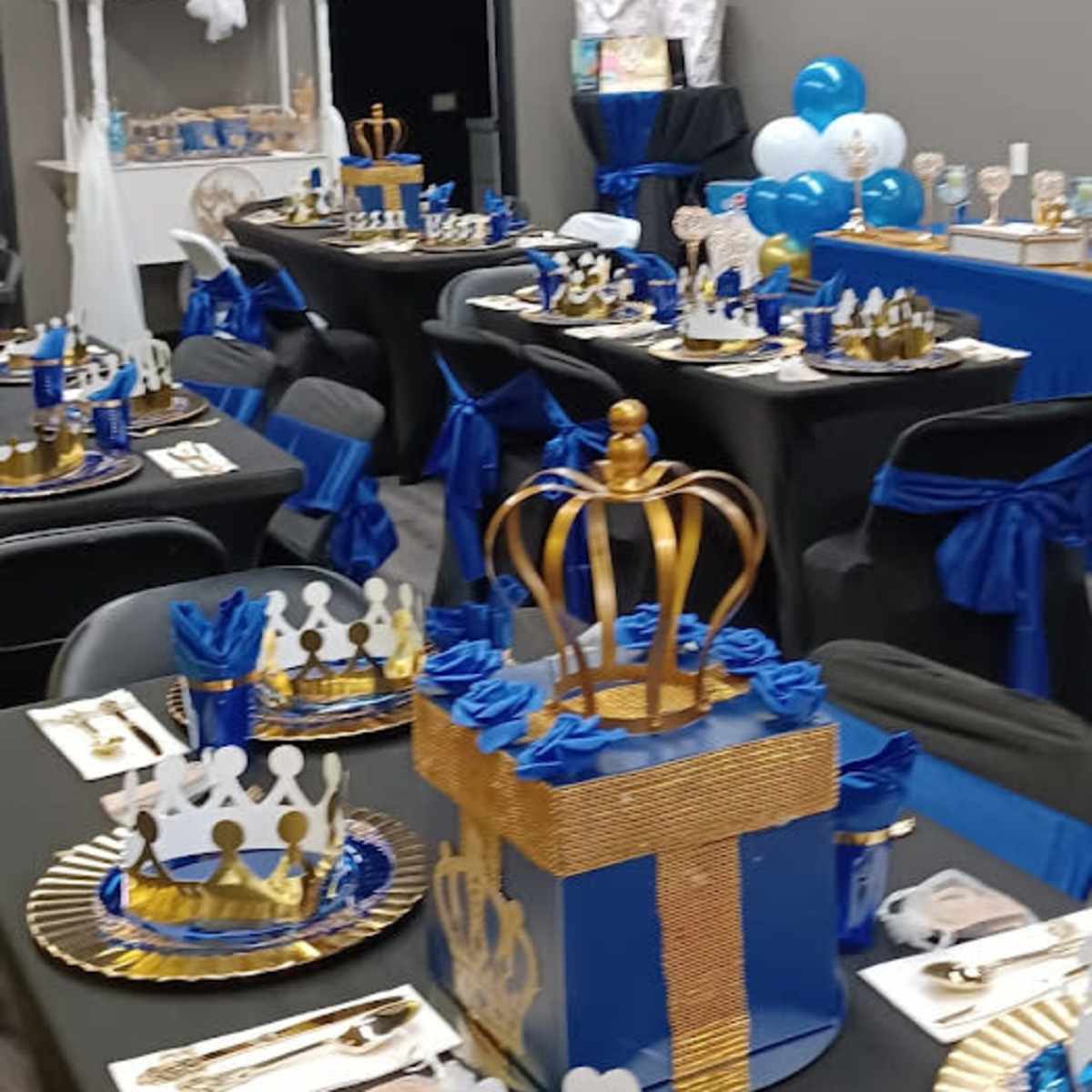 A decorated room with tables set for an event, featuring blue and gold tableware, decorative crowns, and balloons.