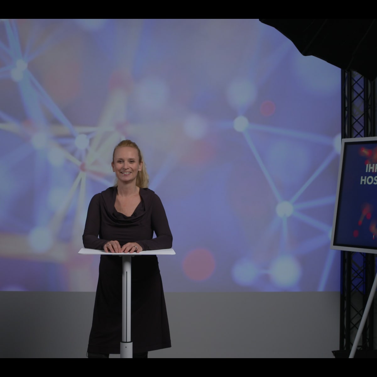 A woman stands behind a podium in front of a colorful, abstract background featuring interconnected lines and dots.