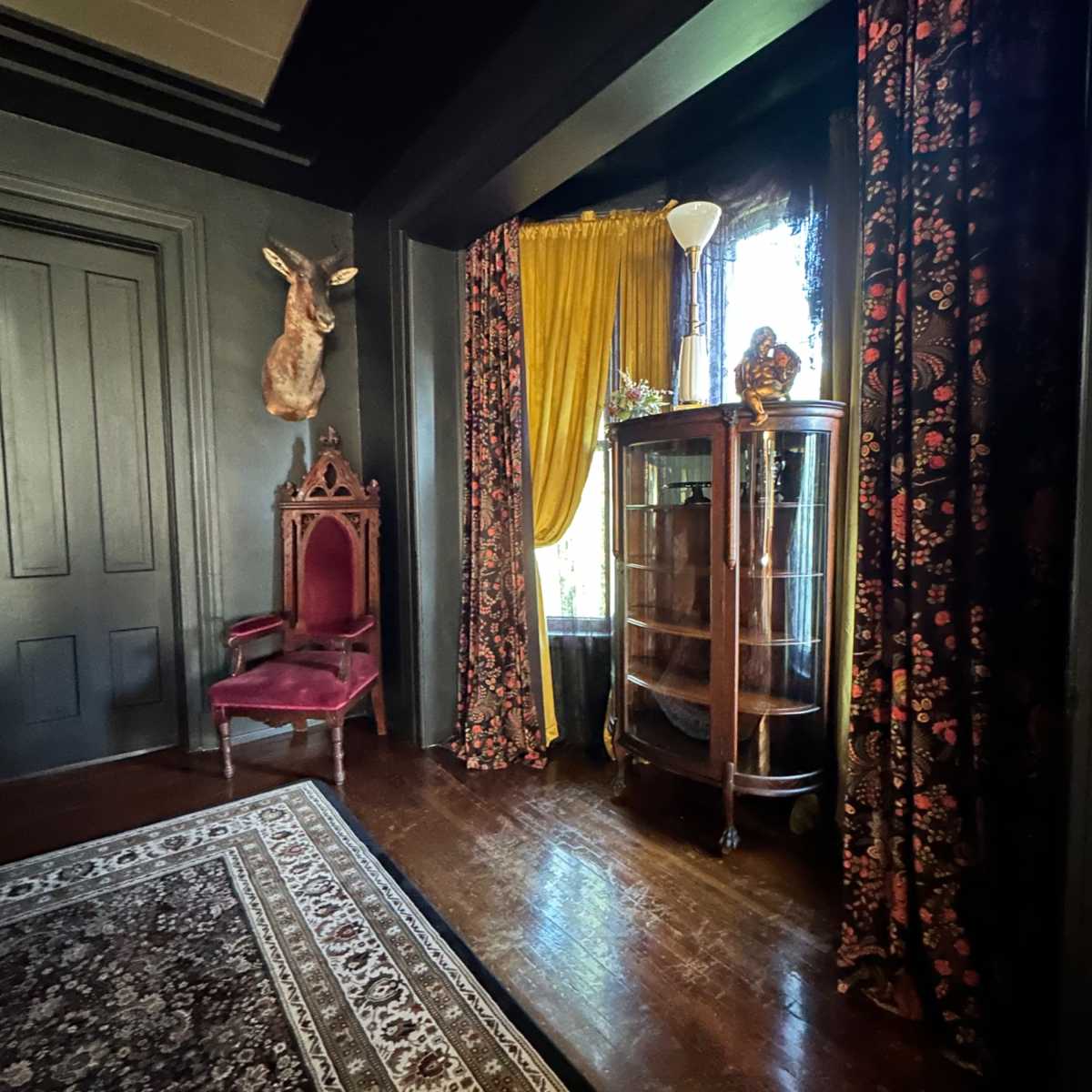 The room features dark walls, a patterned rug, a red chair, a glass display cabinet, and large windows with golden drapes.