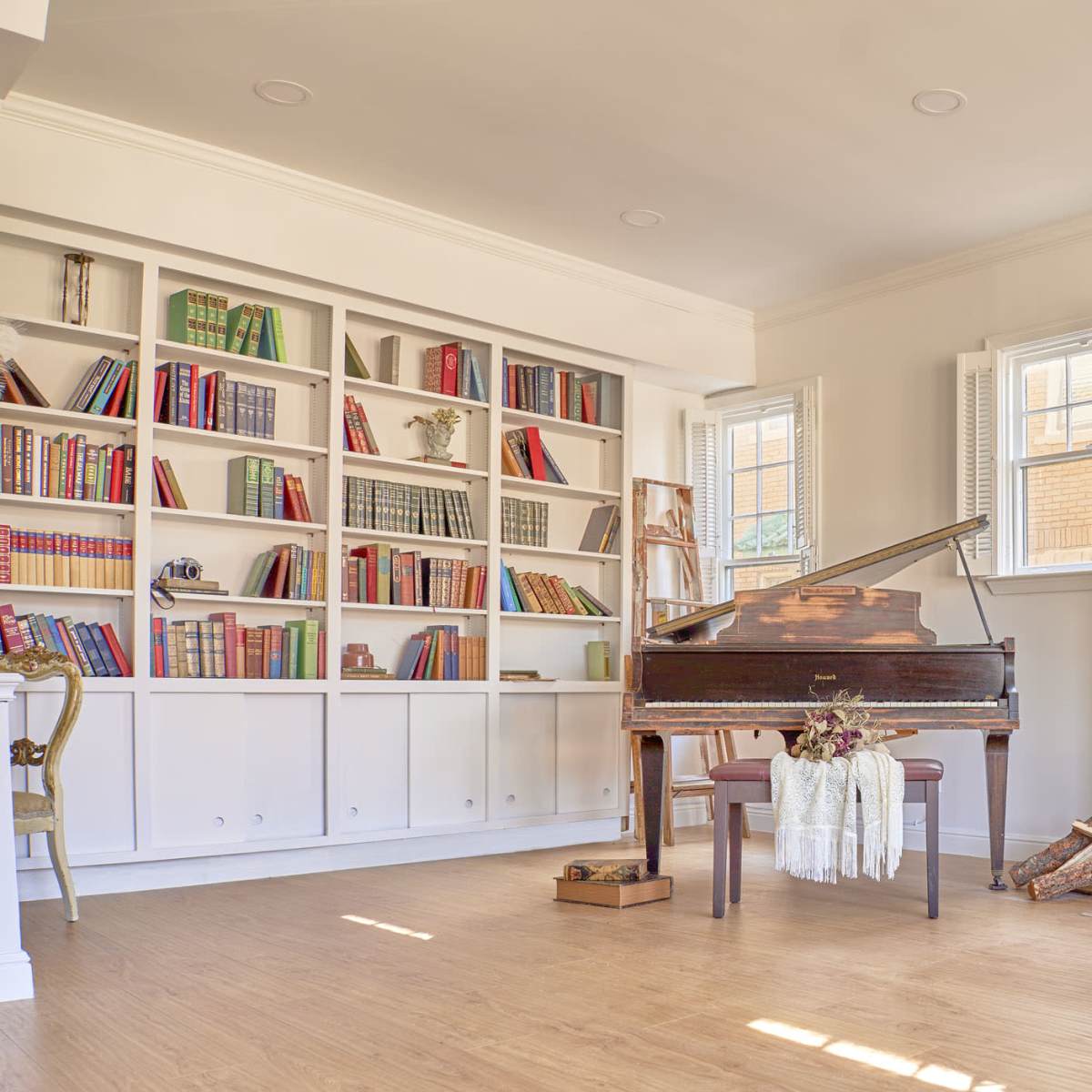 The room features a grand piano in front of a decorative fireplace, with a bookshelf filled with various books and a few decorative items arranged around the space.