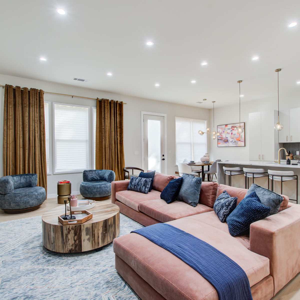 A modern living room with a pink sectional sofa, blue accent chairs, a round wooden coffee table, and a kitchen area in the background featuring white cabinets and pendant lighting.