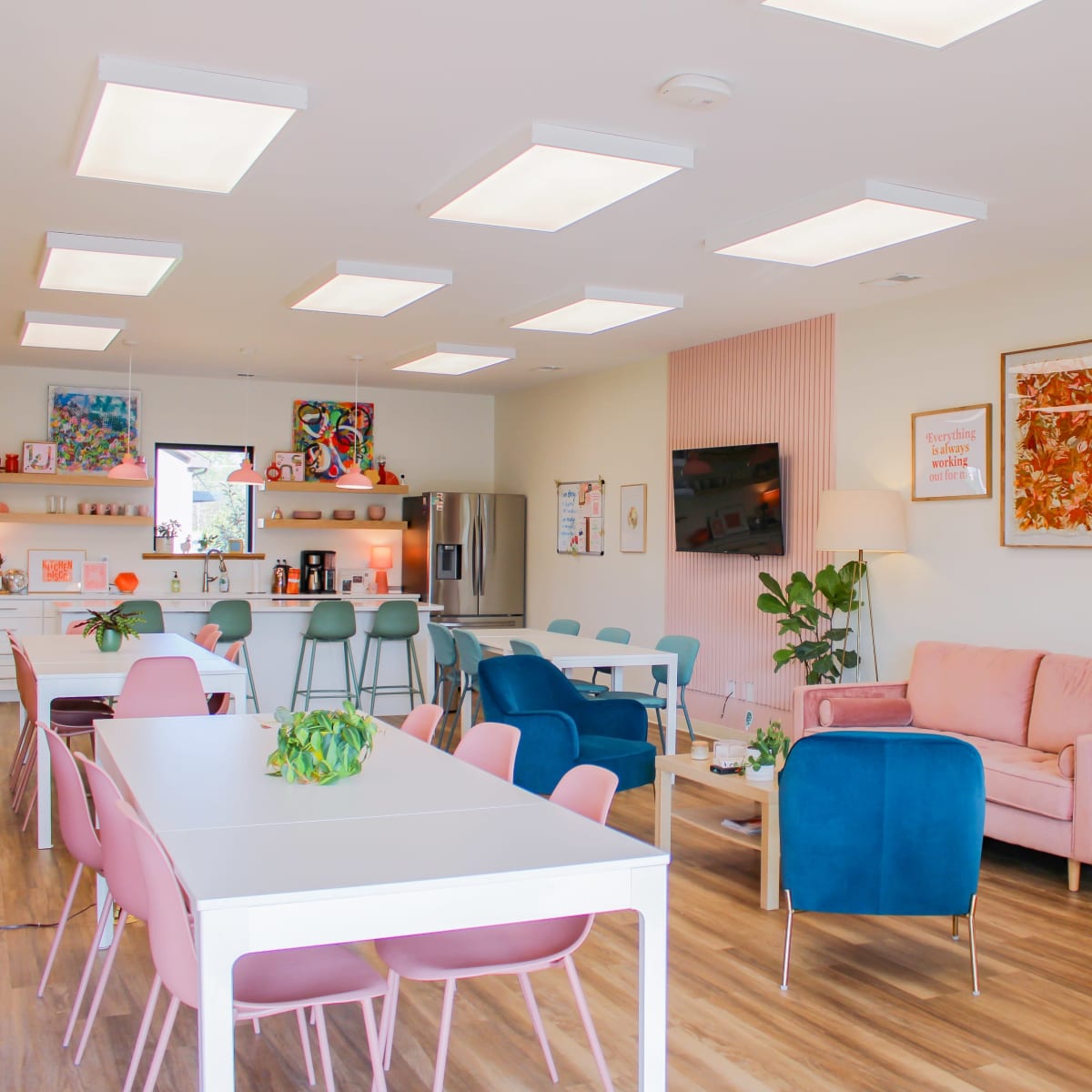 A bright, modern communal space featuring several tables, colorful seating, and a kitchen area in the background.
