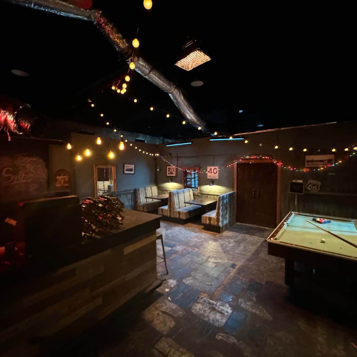 A dimly lit bar interior with a pool table, booths along the walls, and string lights decorating the ceiling.