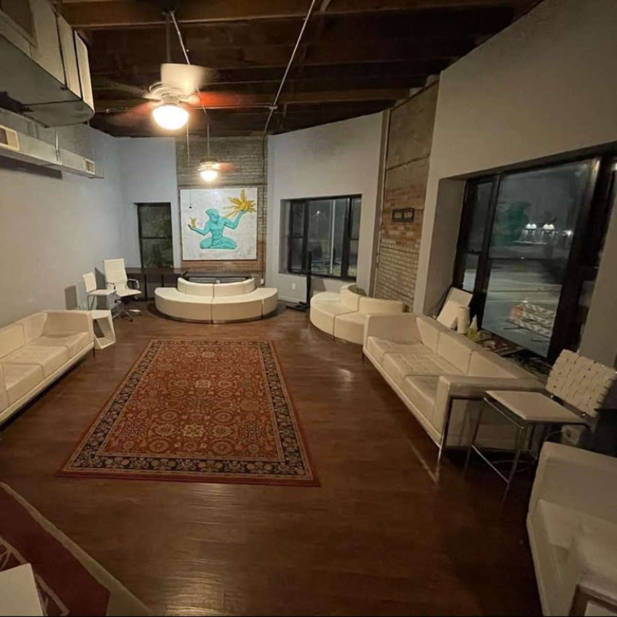 A spacious room with white furniture arranged around a central circular table, featuring a patterned rug on the wooden floor and a large artwork on the wall.
