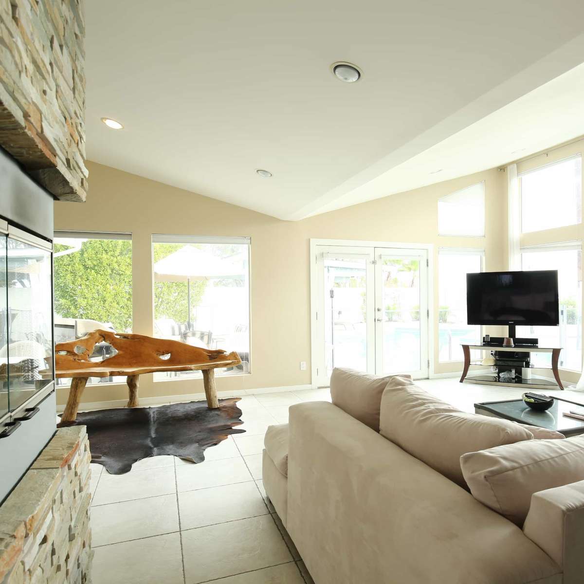 A modern living room with a stone fireplace, a large sofa, and floor-to-ceiling windows overlooking a pool area.