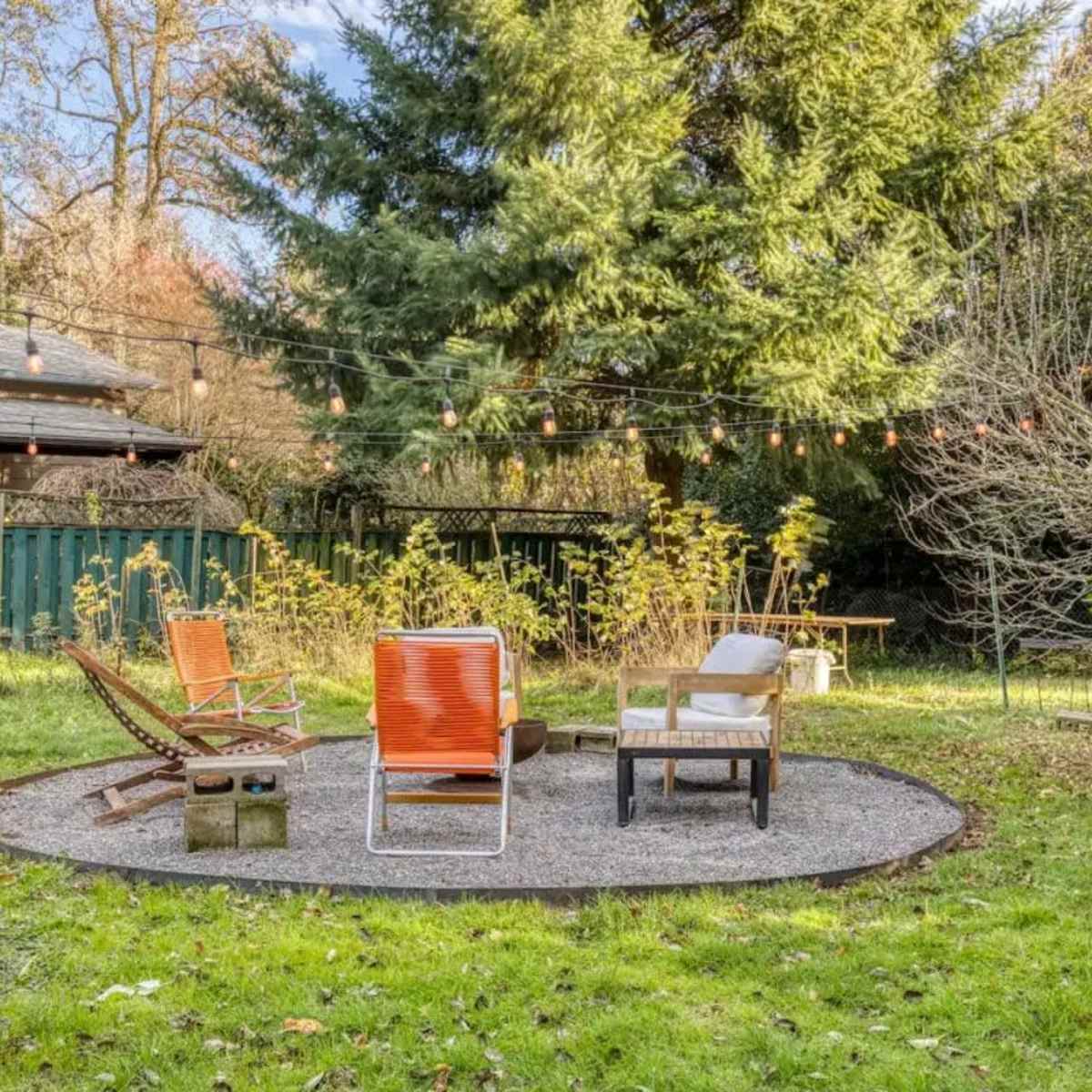 A circular gravel sitting area features orange chairs and a small table, surrounded by greenery and string lights in a backyard.