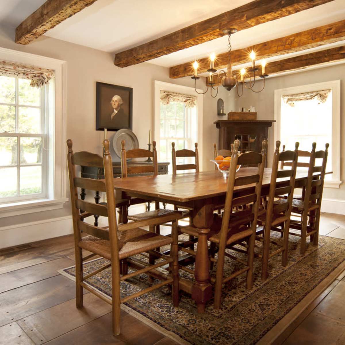 A rustic dining room with a large wooden table surrounded by ten chairs, illuminated by a chandelier and featuring exposed wooden beams and a fireplace.