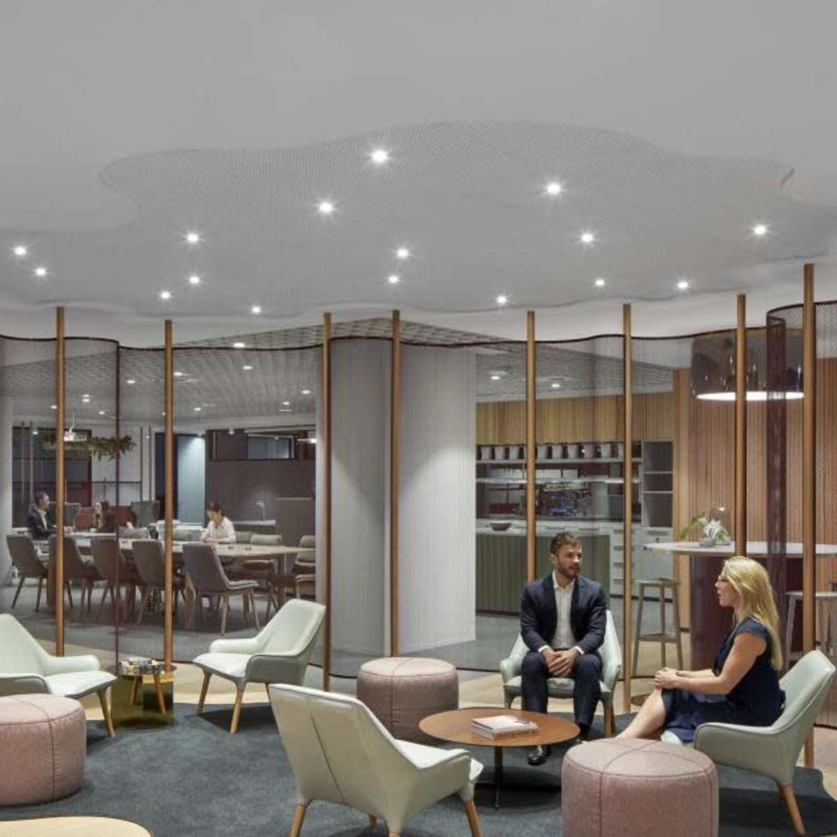 A modern office lobby features seating arrangements around a circular rug, with people engaged in conversation and a backdrop of wooden elements and plants.