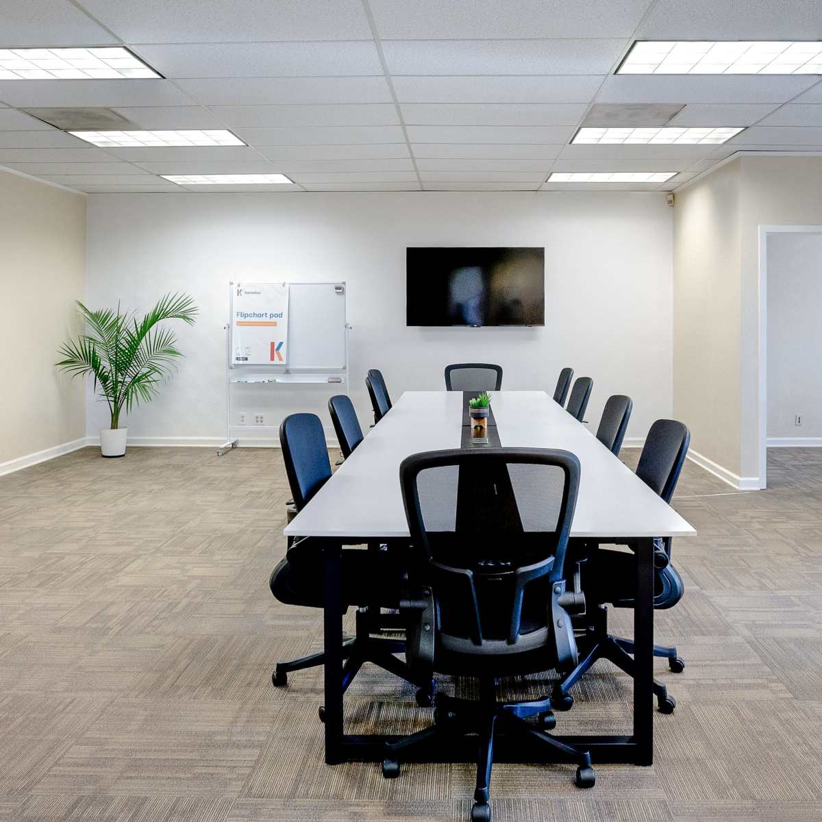 A conference room features a long table with chairs, a television mounted on the wall, and a whiteboard, complemented by a plant in the corner.