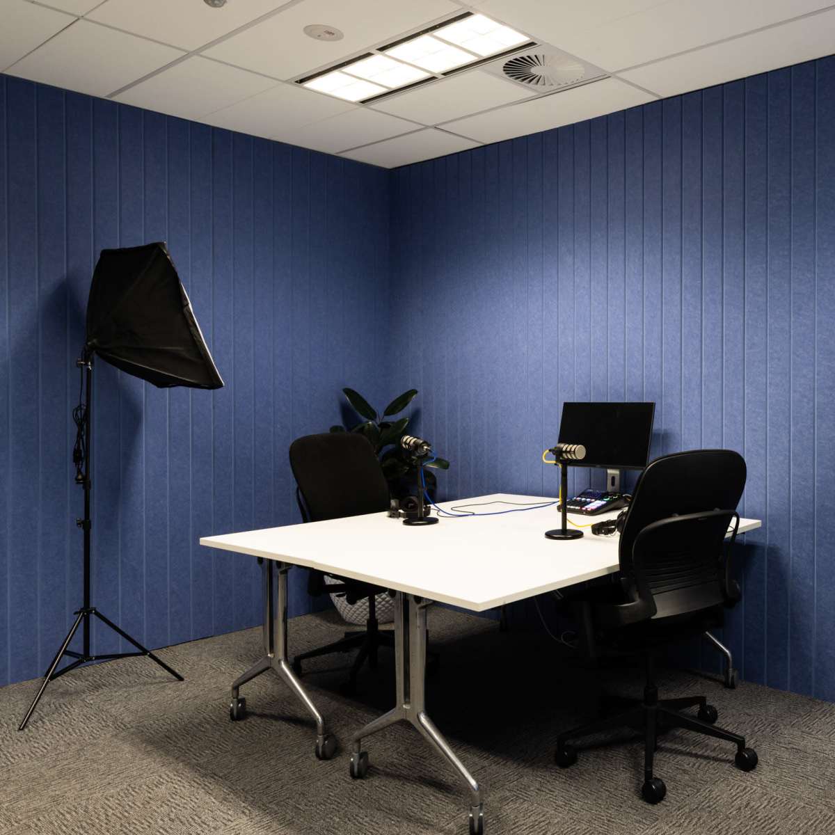 A small office is set up for video recording, featuring a white table, two black chairs, and professional lighting equipment against blue walls.