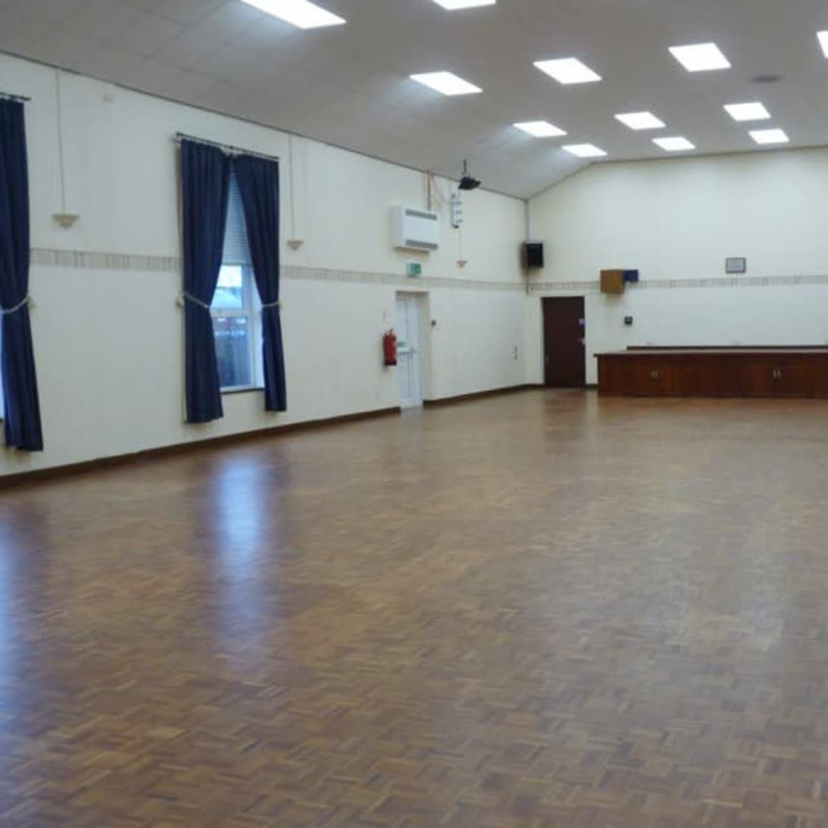 A large, empty hall with wooden flooring, ceiling lights, and a service counter at the far end.