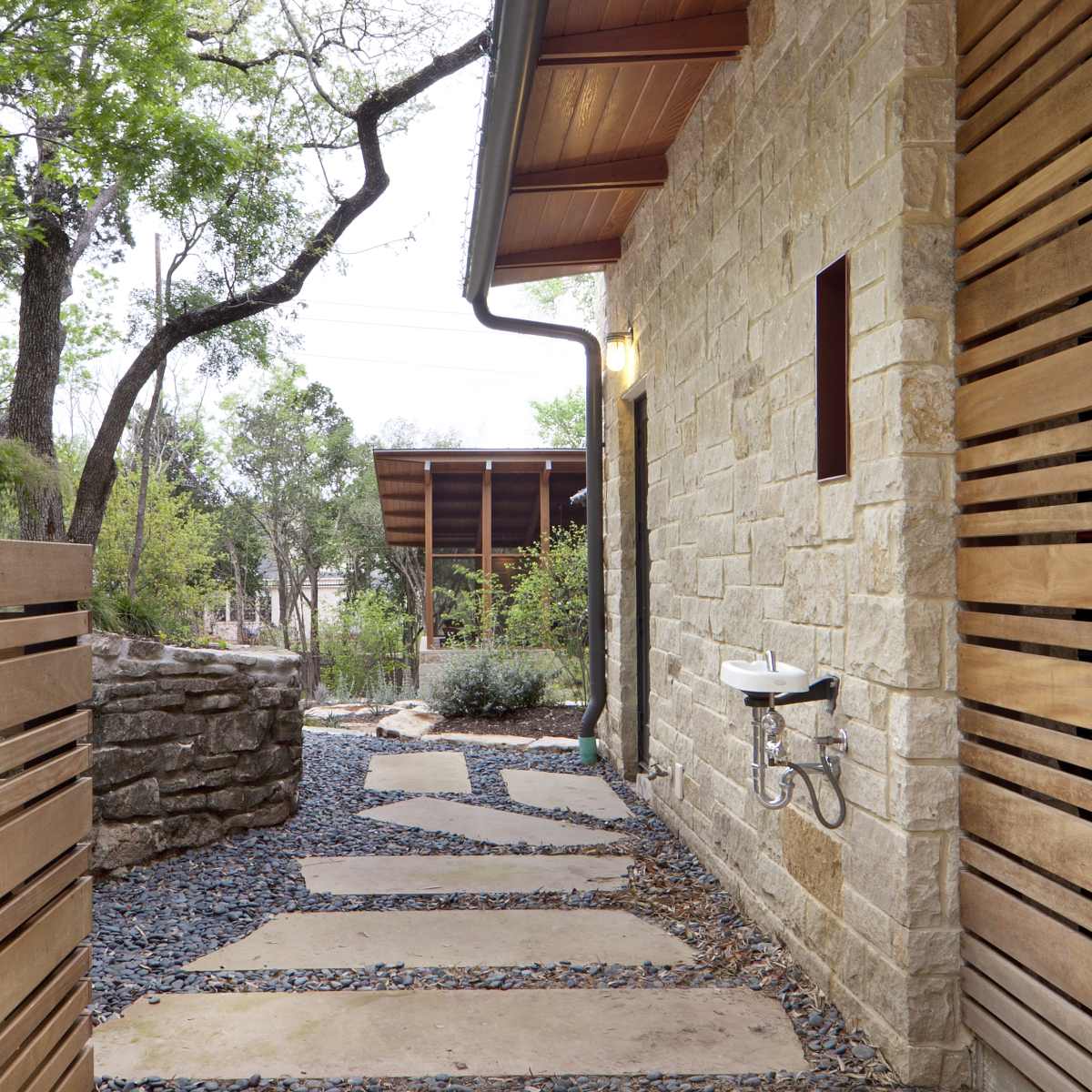 A stone pathway bordered by landscaped greenery, leading toward a building with wooden siding and a covered area in the background.