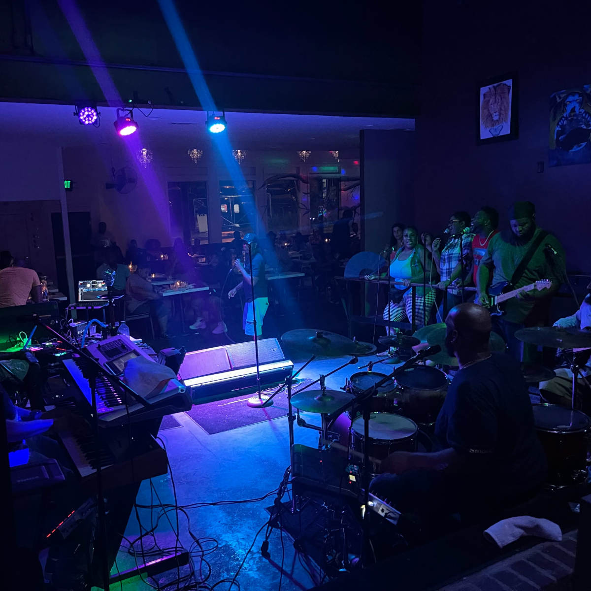 A band performs on stage in a dimly lit venue while a small audience watches from the side.