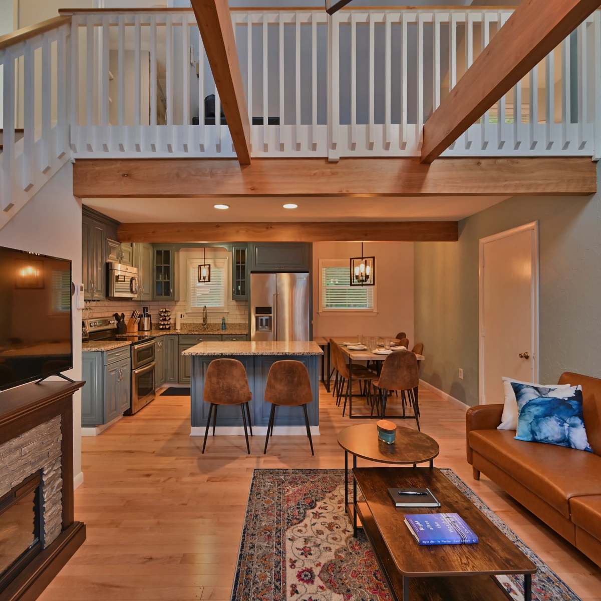 A modern living space with a staircase, an open kitchen area, a living room featuring a leather couch, and wooden beams across the ceiling.