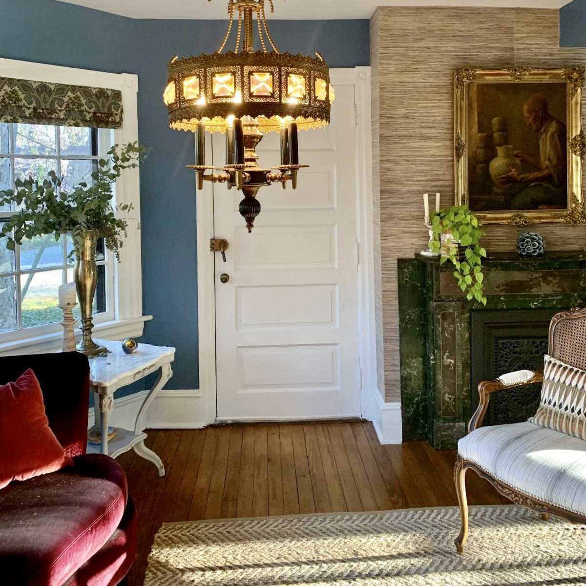 A decorative chandelier hangs from the ceiling in a room with blue walls and hardwood flooring, featuring a white door and a fireplace with a painting above it.