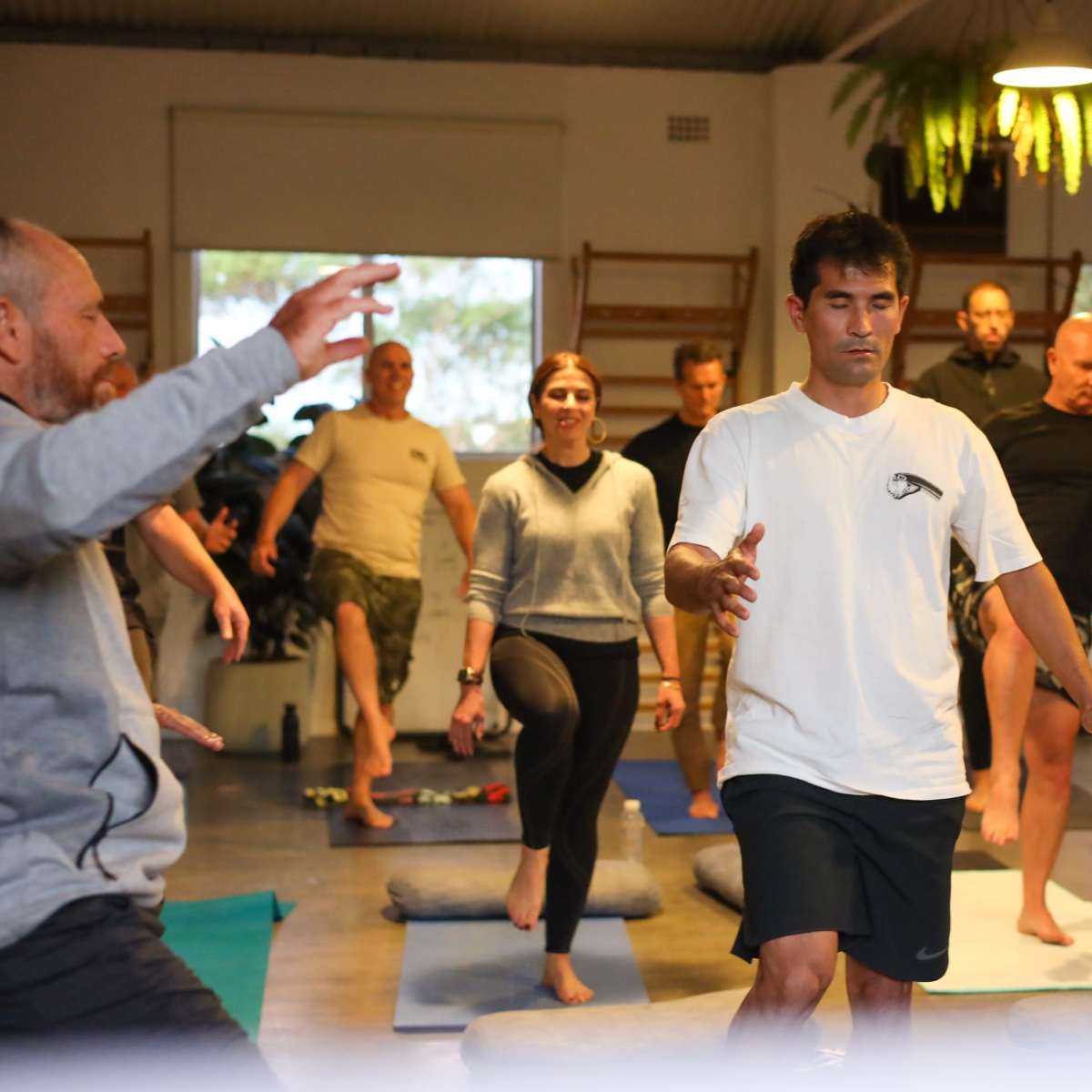 A group of people practice yoga in a spacious studio with mats and cushions on the floor.