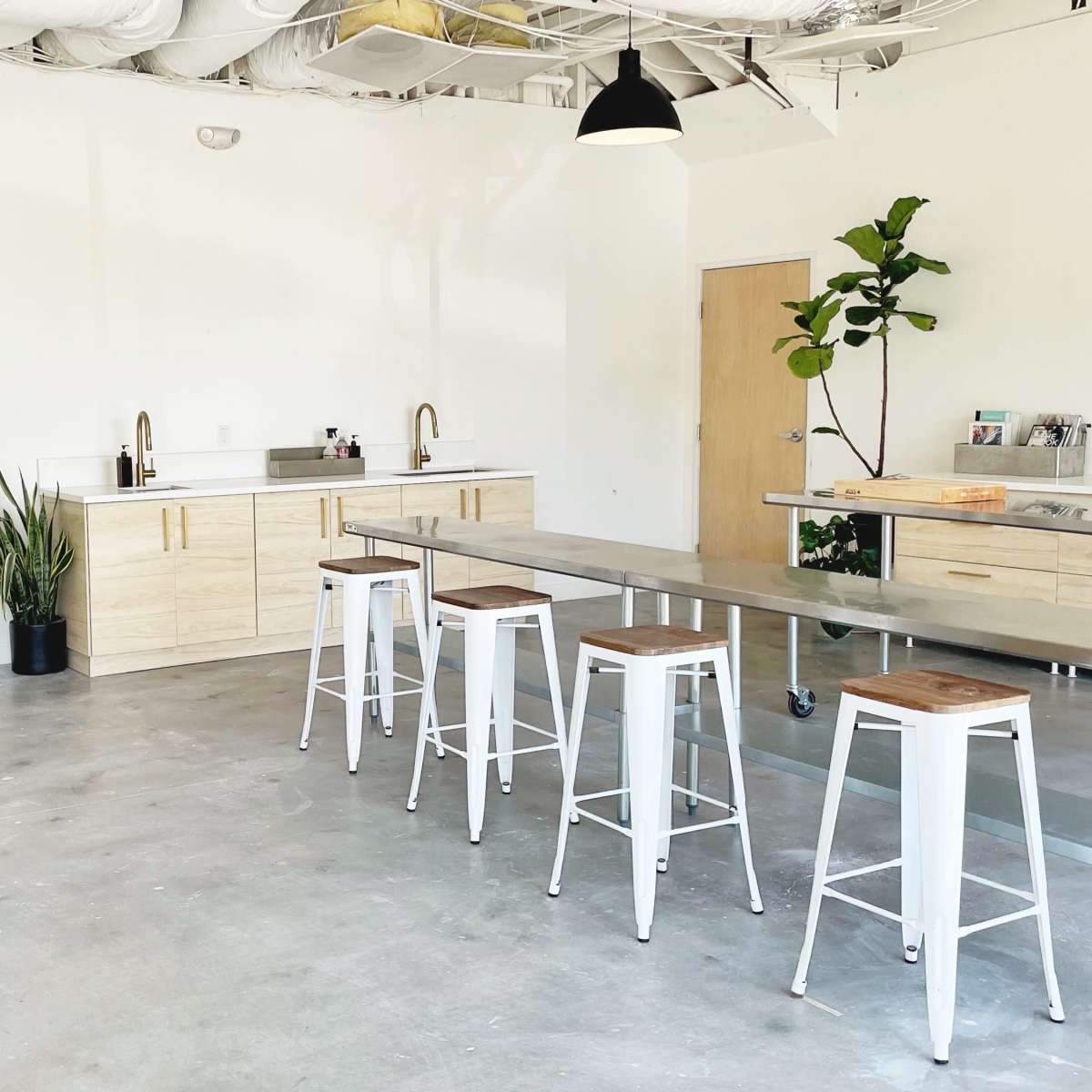 A spacious room features a kitchen area with wooden cabinetry, a metal table with stools, and two wire chairs, along with a plant in the corner.