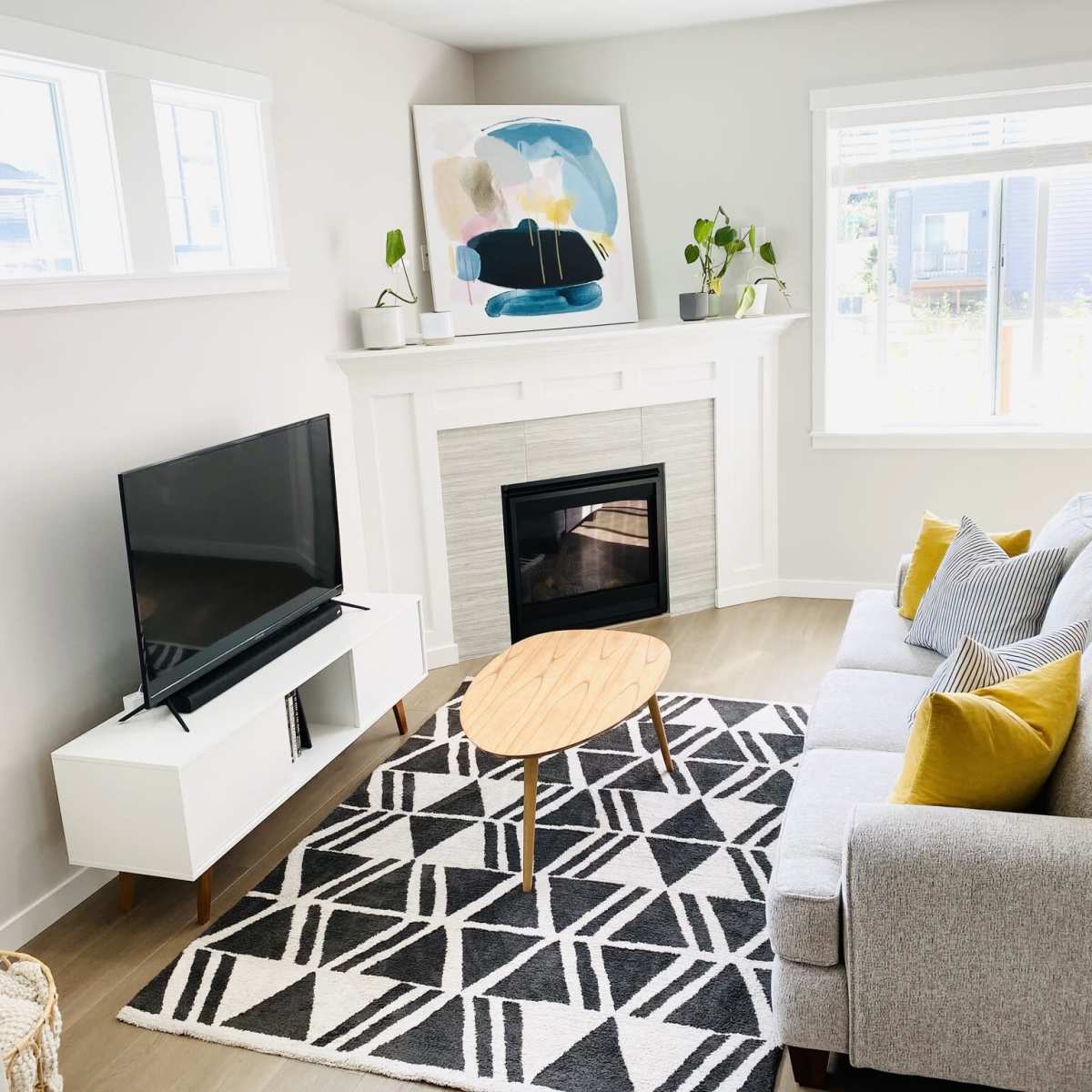 A modern living room features a gray sofa, a wooden coffee table, a television on a white media console, and a fireplace beneath a large artwork, with natural light coming through the windows.