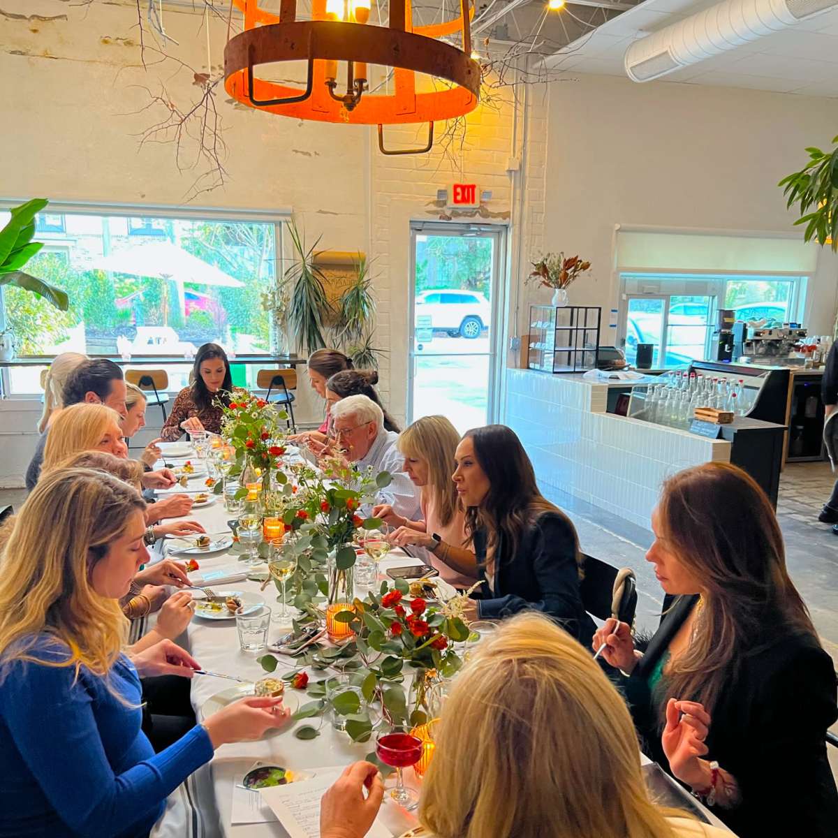 A group of people is seated at a long table adorned with flowers, enjoying a meal in a brightly lit restaurant.