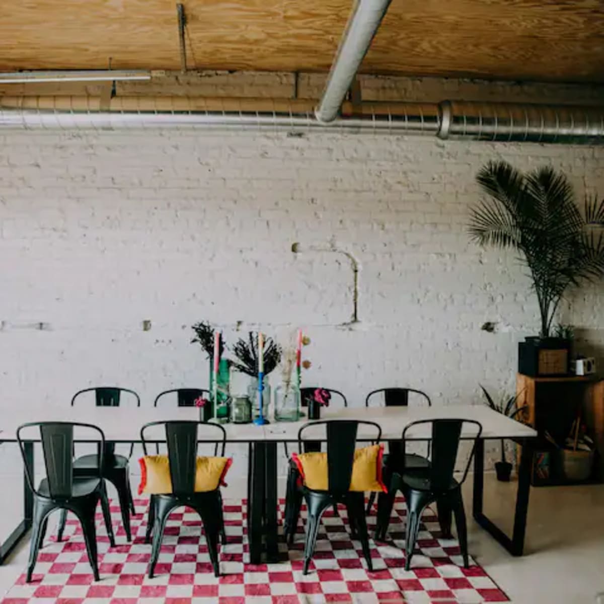 A long dining table surrounded by black chairs is set against a white brick wall, with colorful decorations and a patterned rug underneath.