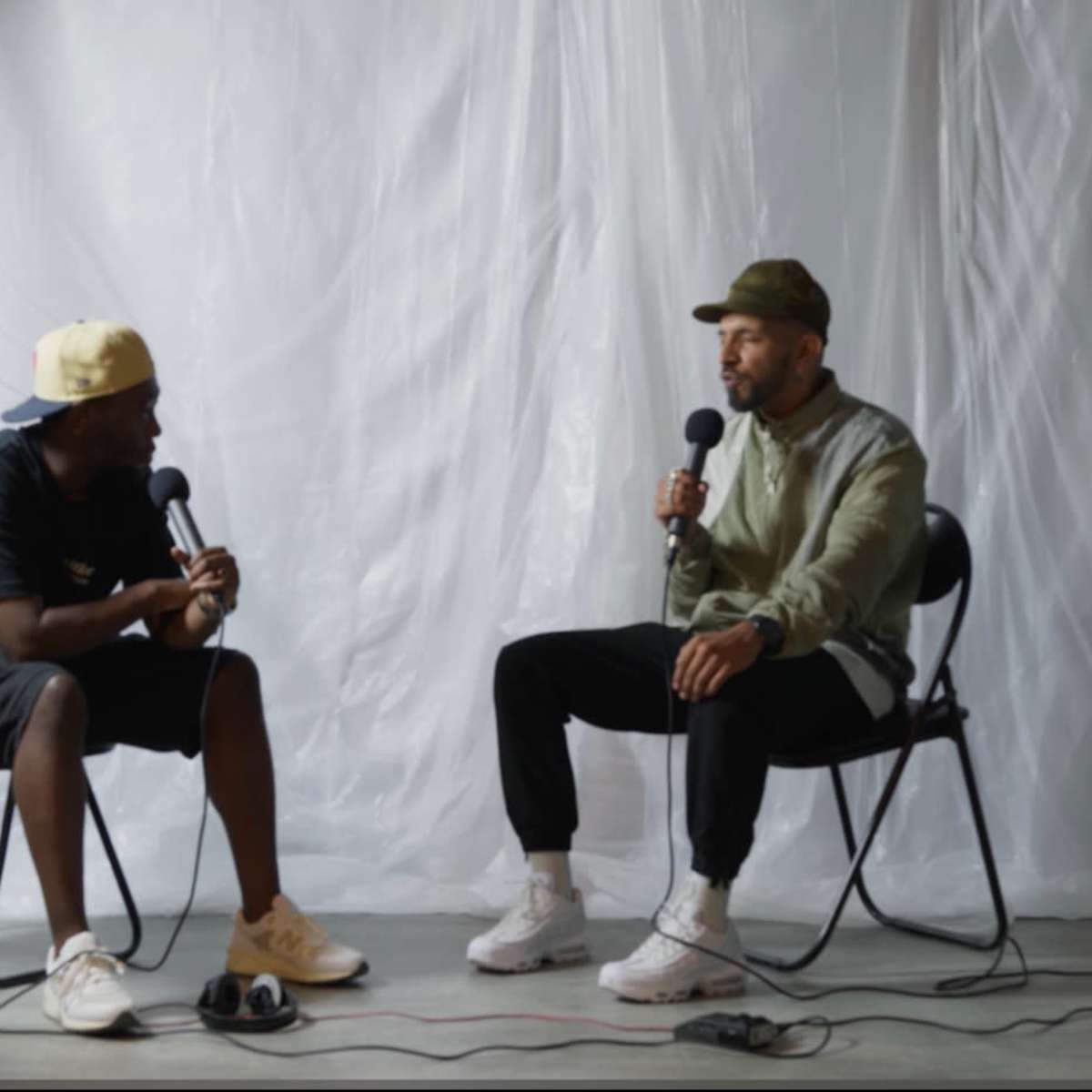 Two men sit in separate chairs, engaged in a conversation while holding microphones, against a white backdrop.