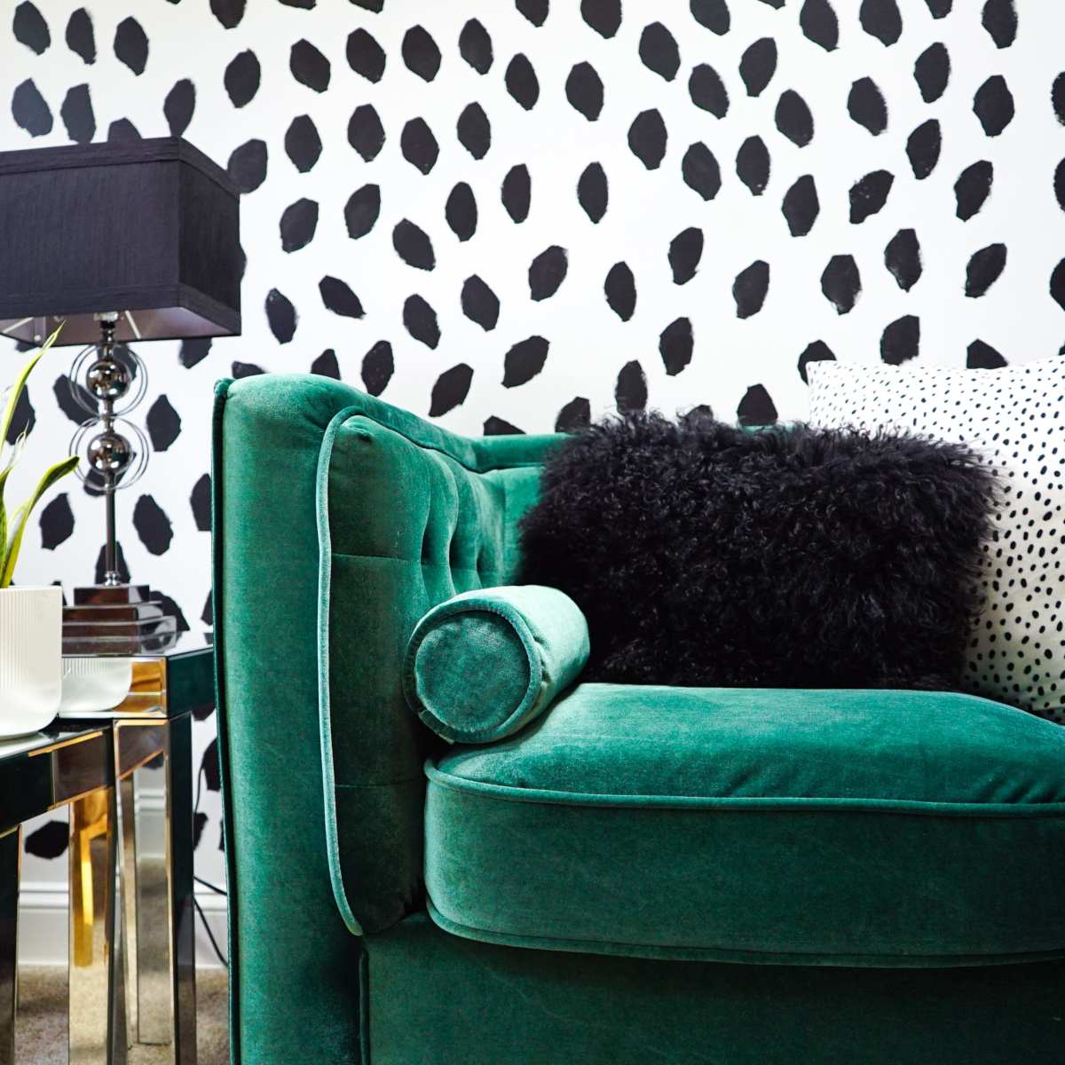 A green velvet sofa adorned with various decorative pillows, next to a stylish side table with a lamp and a potted plant, all against a black-and-white dotted wall.