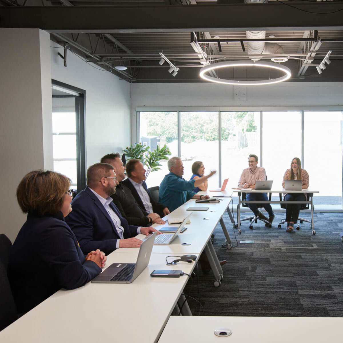A group of eight professionals is seated at a long table in a modern conference room, while a woman stands at the front presenting to the team.
