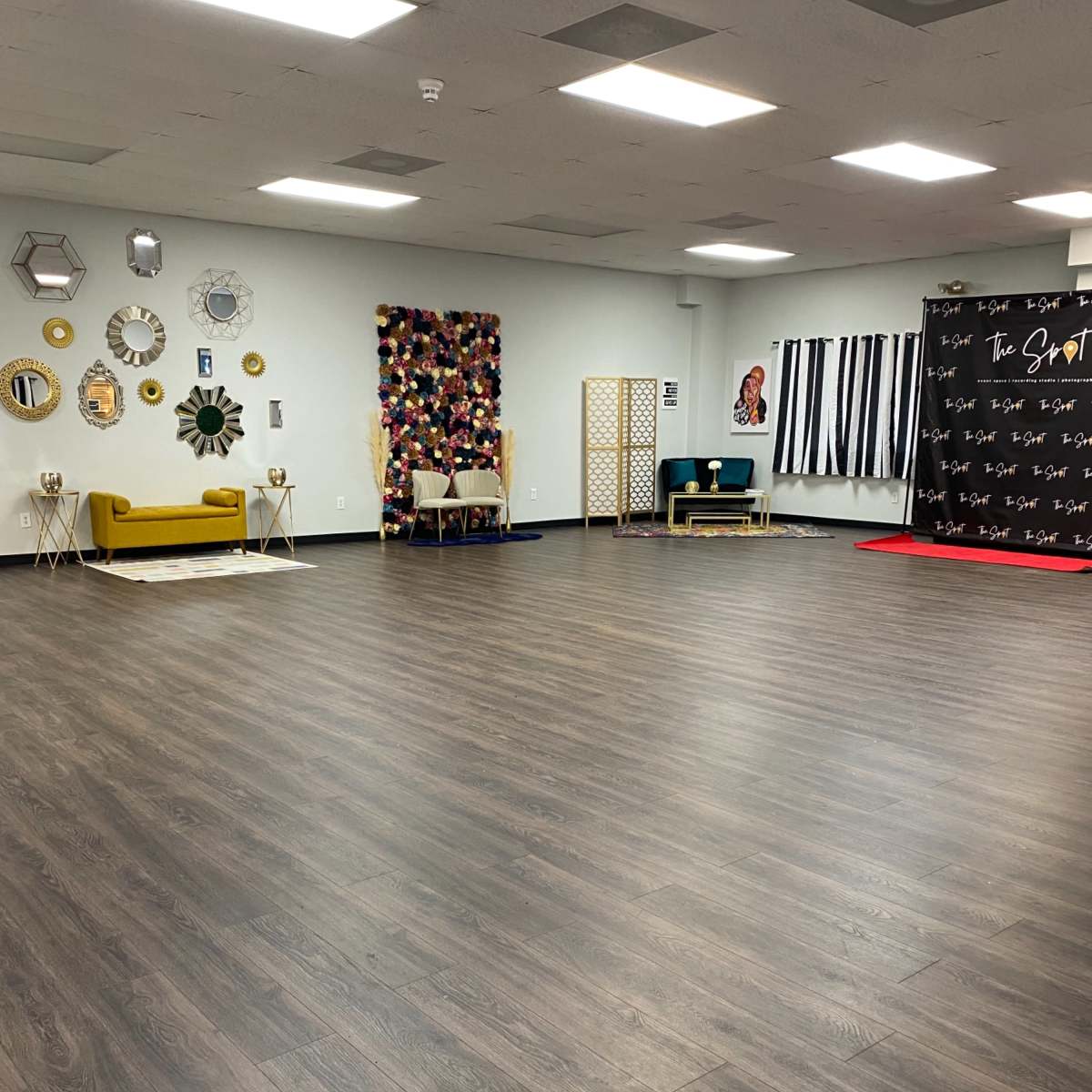 A spacious, empty room with wooden flooring, decorated walls featuring various wall hangings, seating areas, and a backdrop for events.