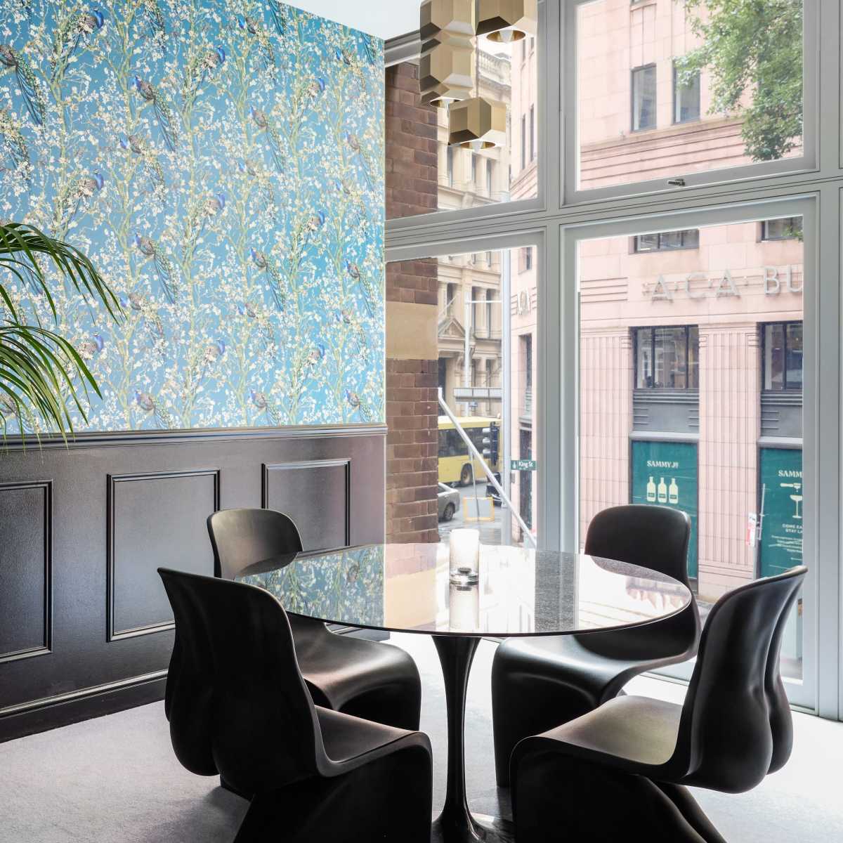A modern dining area features four black chairs around a round glass table, set against a blue floral wallpaper and large windows with a city view.