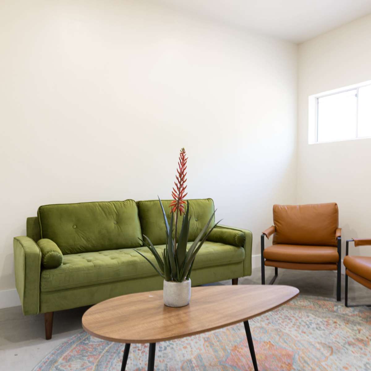 The room features a green velvet sofa, two brown leather chairs, a round wooden coffee table, and a potted plant on the table, with a small window providing natural light.