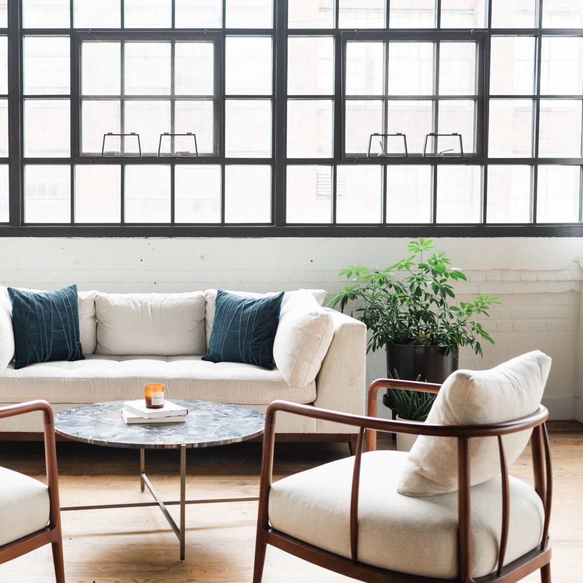 A light-colored sectional sofa paired with two wooden chairs and a round marble coffee table sits in front of large industrial windows.
