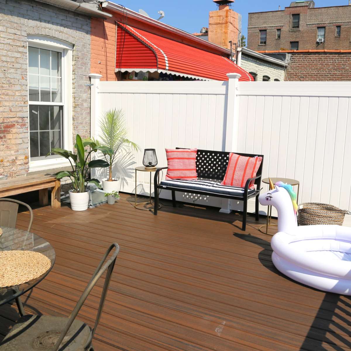 A wooden deck area featuring a table with chairs, a bench with an American flag cushion, potted plants, and a large inflatable unicorn.