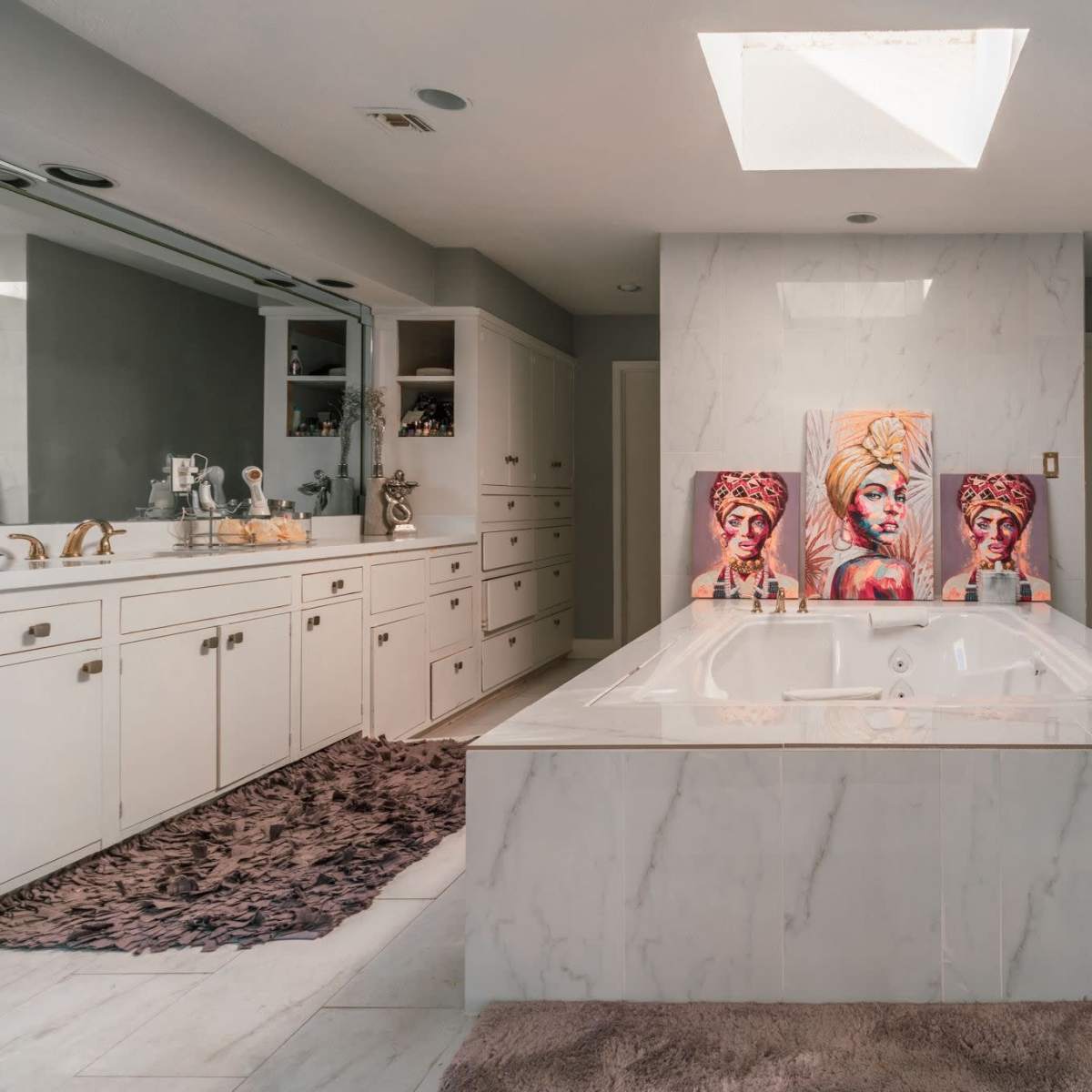A spacious bathroom features a central white marble bathtub, double vanities with storage cabinets, and artwork of women adorning the walls.