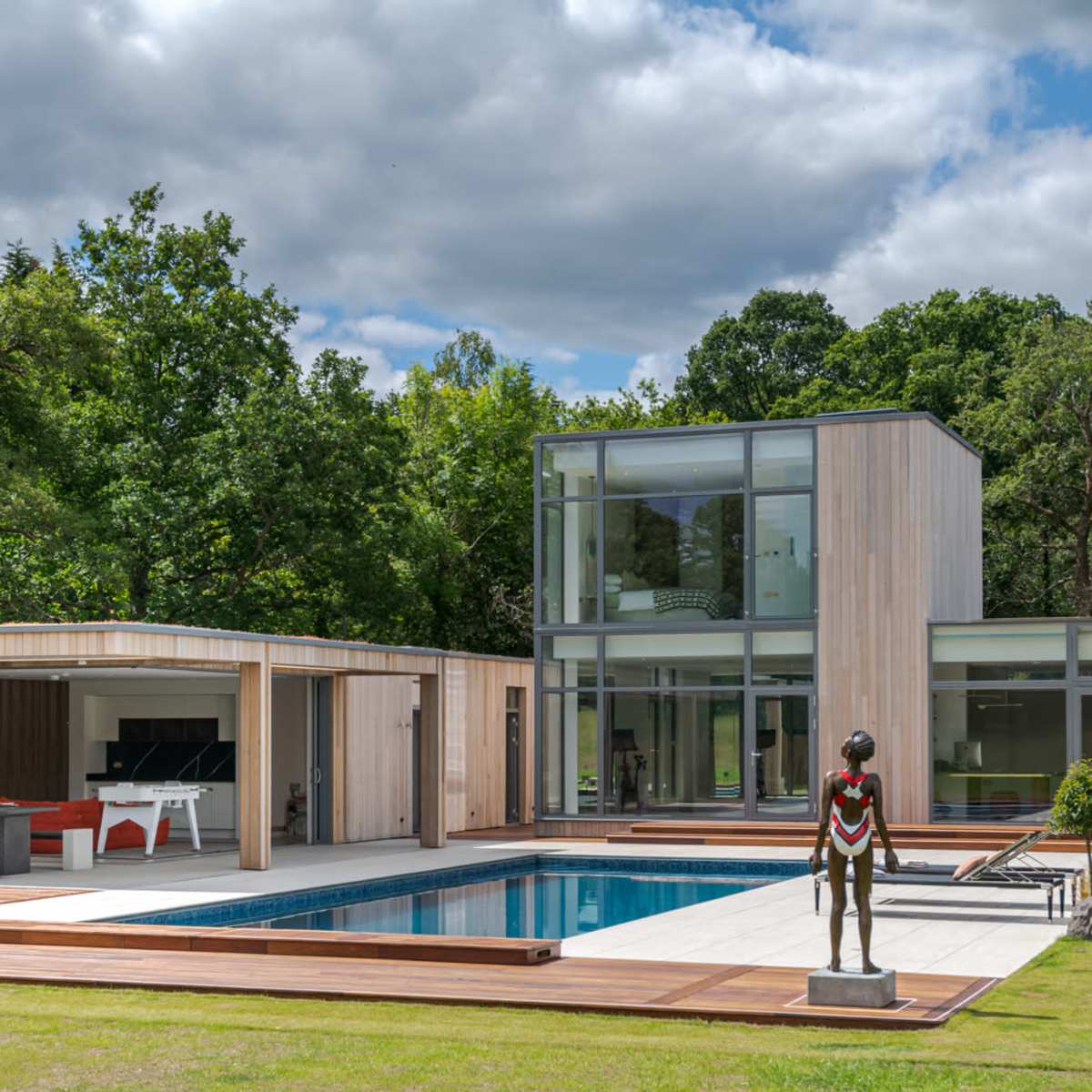 A modern house with large glass windows, a swimming pool, and a sculpture in the foreground, all set in a landscaped area with trees.