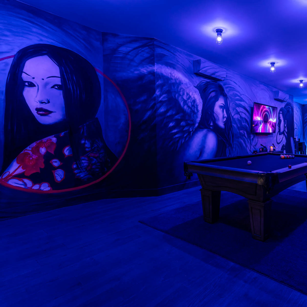 A dimly lit room featuring a pool table surrounded by murals of women with an artistic and colorful design.