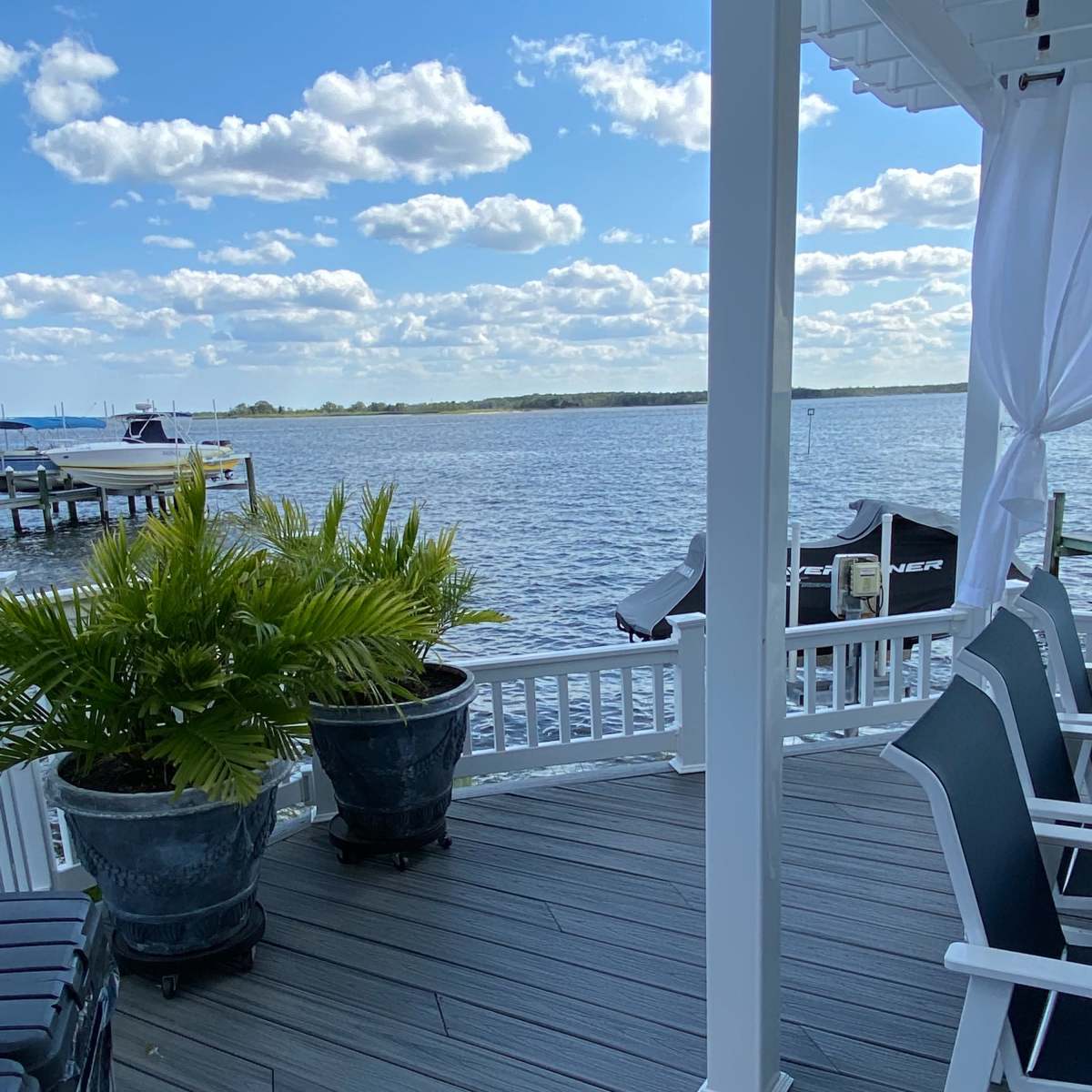 A riverfront deck area with two potted ferns and black chairs overlooking the water.