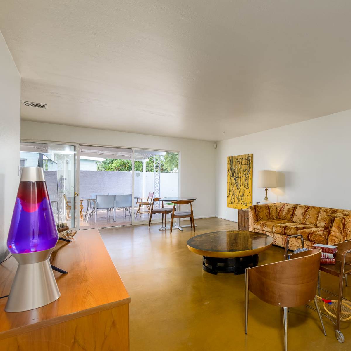 A living room with a brown sofa, a round coffee table, and a lava lamp, with large windows revealing an outdoor patio area.
