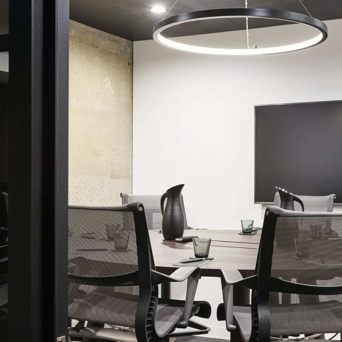 A conference room features a round table surrounded by several mesh chairs, with a large screen mounted on the wall and a door labeled "Meeting Room."